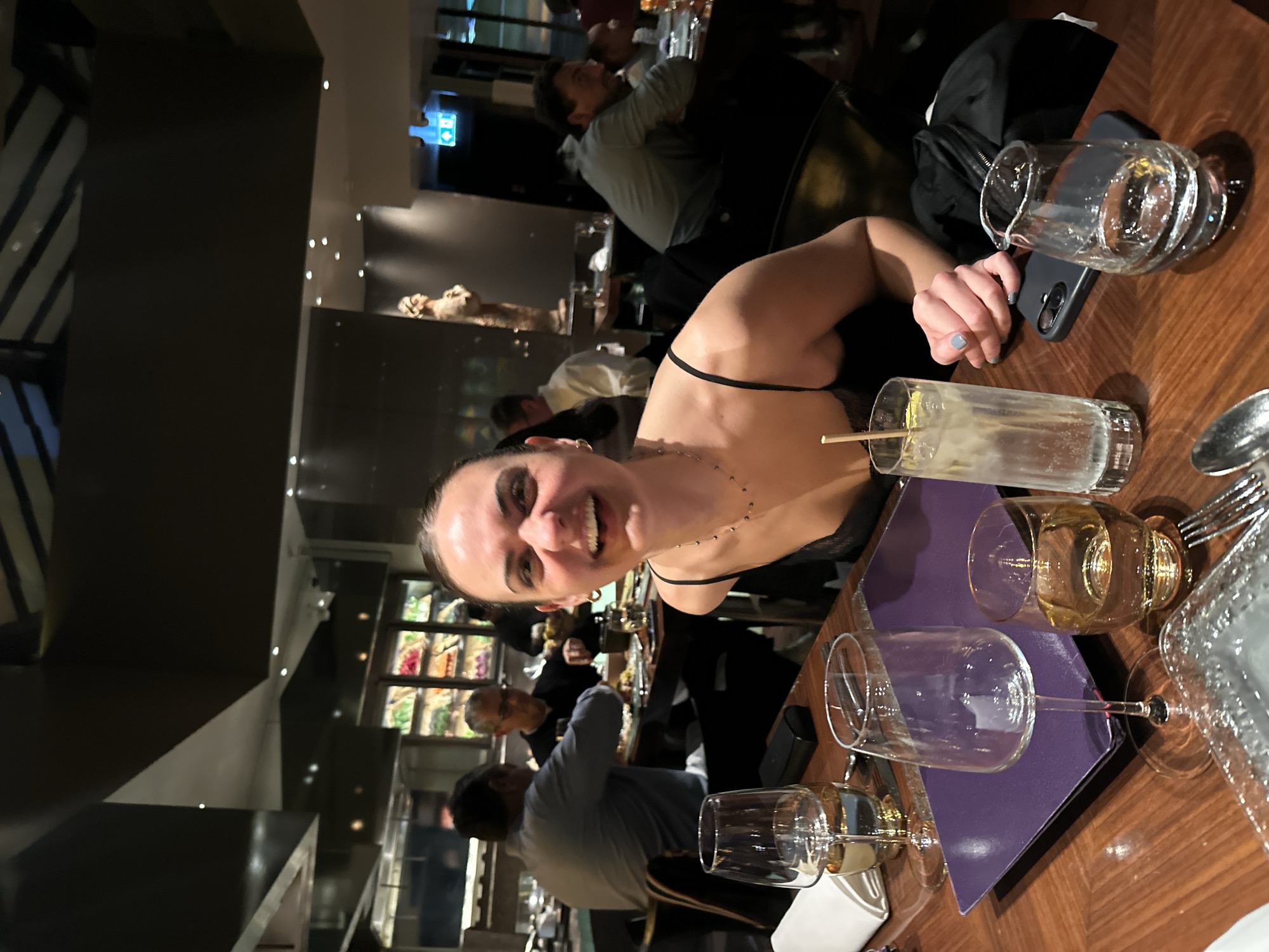 The image shows a woman smiling at the camera while seated at a restaurant table in what appears to be an upmarket dining establishment. The restaurant appears lively with other patrons visible in the background. The table is set with glassware, including wine glasses and water jugs. A mobile phone is also on the table. The lighting is ambient. The precise location cannot be definitively determined but given the overall aesthetic and the presence of a multicultural clientele, it is plausible that this scene is taking place in a restaurant within the city of London, United Kingdom.
