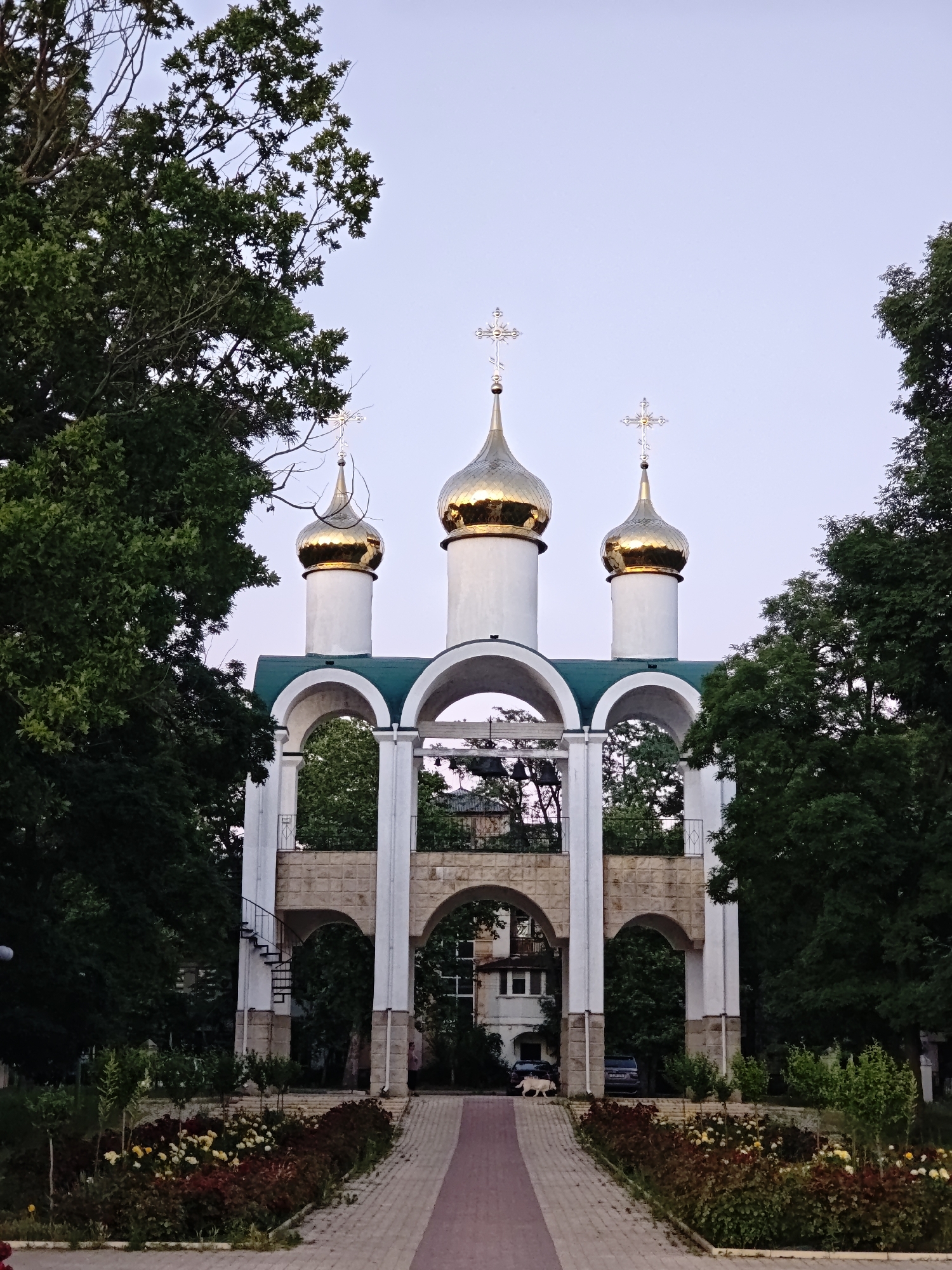 An exterior, low-angle, vertical shot captures an ornate church bell tower with three golden domes topped with crosses. The tower is flanked by lush green trees and is adorned with white arches and stone. In the foreground, a pathway leads to the bell tower, bordered by flower beds with red and yellow blossoms. Two vehicles and a white dog are visible near the base of the bell tower. The sky above is a pale, clear blue. This appears to be the bell tower of the Nativity Cathedral in Tiraspol, Moldova.