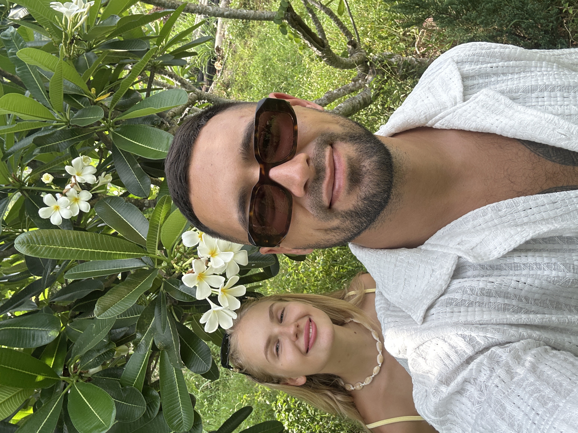 This media file is a portrait of a couple outdoors in a tropical setting. The man is in the foreground, wearing sunglasses and a white robe. He has dark hair and a beard. The woman is behind him, smiling and looking at the camera. She has blonde hair and is wearing a yellow top and a shell necklace.
They are surrounded by lush greenery, including a tree with large green leaves and white flowers, possibly frangipani. The background consists of more greenery and dappled sunlight, suggesting it is daytime. The overall mood is relaxed and happy. The presence of tropical flora strongly suggests a warm climate and likely a vacation setting.
The image is a selfie taken at eye level, with the subjects looking directly at the camera. The orientation of the image is such that the subjects appear upside down relative to a typical upright portrait. There is no visible text in the image. The location context provided is Nam Nha Trang Ward, Vietnam, which aligns with the visual cues of a tropical environment.