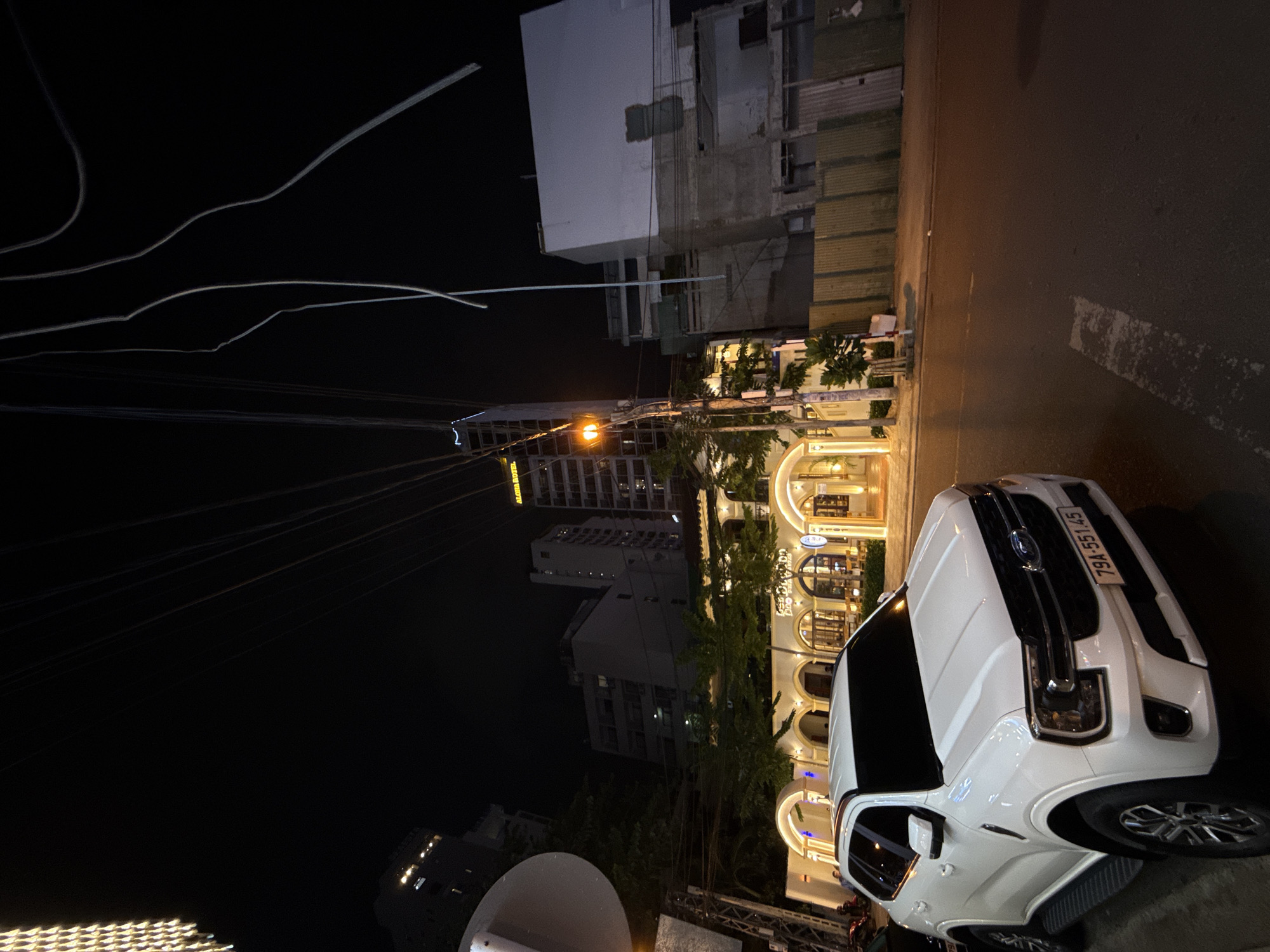 In Nha Trang, Vietnam, at night, a white Ford Everest SUV is parked on the side of a street. The vehicle faces right, displaying its front bumper and license plate, which reads "79A-551.45". Adjacent to the street, illuminated buildings with arched entrances and warm lighting suggest a commercial area, possibly restaurants or shops. Trees are interspersed along the facade of these buildings. In the background, taller buildings with illuminated windows rise into the dark sky, indicating an urban setting. Overhead, a tangle of power lines is visible against the night sky, some appearing damaged or disconnected. The street surface is asphalt with painted white lane markings. The overall atmosphere is one of an active city street at night, with the parked SUV as a prominent subject.