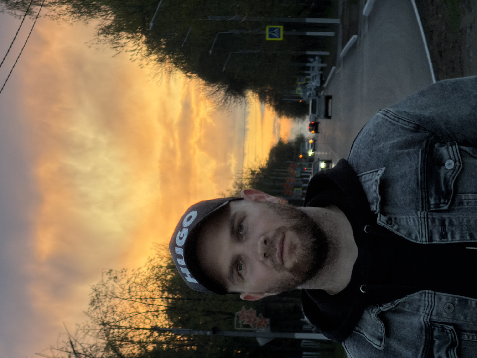 A man is positioned in the foreground of an image with a street scene and sunset backdrop. The man has a beard and is wearing a dark "HUGO" baseball cap, a black hoodie, and a gray denim jacket. He is looking directly at the camera.
Behind him, there's a road with multiple vehicles, including cars and trucks, visible on it. Street lights and foliage line the sides of the road. Traffic signs, including a pedestrian crossing sign, are also visible.
The sky behind the street scene features a vibrant sunset, with colors ranging from yellow to orange and gray. It appears to be in the city of Chaikovsky, Russia, according to the prompt.