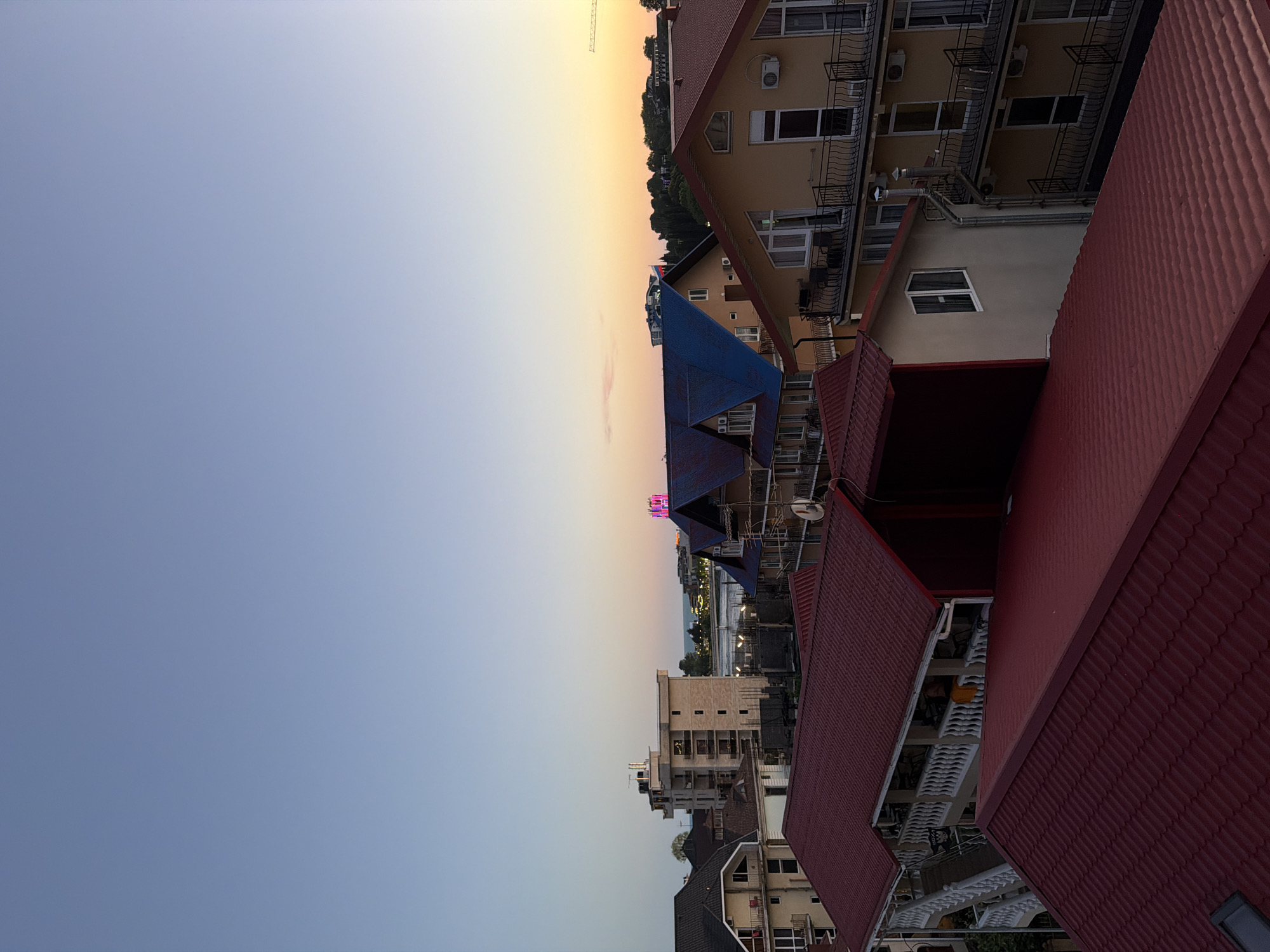 An aerial, high-angle shot captures a twilight view of buildings with red and blue roofs in Sirius, Russia. The sky transitions from a clear, pale blue at the top to a soft orange and pink hue near the horizon, suggesting dusk. In the middle ground, several multi-story buildings are visible, one with a distinct blue roof. Further in the distance, the sea can be seen, bordered by more buildings and trees. A prominent, bright pink neon sign is illuminated on a building in the far distance. The foreground is dominated by the textured, red tiled roofs of several structures. The scene appears to be a residential or tourist area, given the architecture.