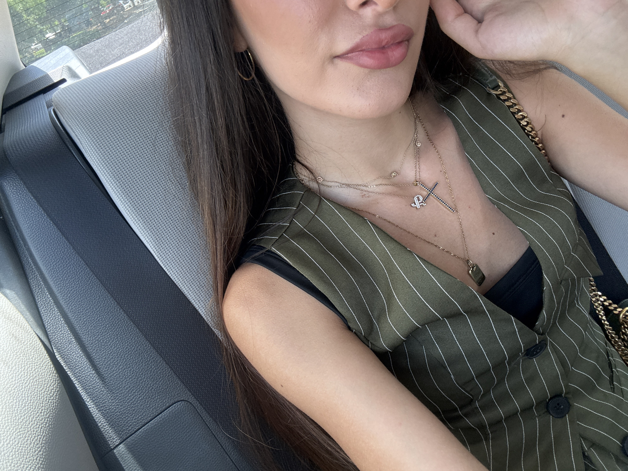 A close-up, overhead shot shows a woman sitting in the back seat of a car. She has long, dark hair and is wearing a green pinstripe vest over a black top and multiple necklaces. The interior of the car appears to be made of dark gray leather and a light-colored fabric. The woman's right hand is resting on her chin. Outside the car window, trees and a hazy sky are visible. This image was taken on November 3, 2023.