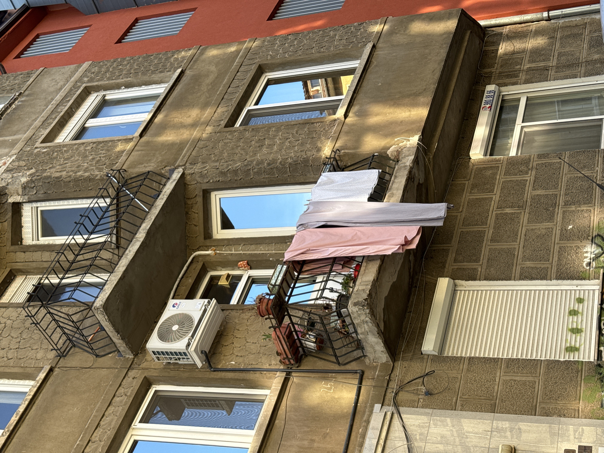 The image captures a portion of a weathered, multi-story building in Sofia, Bulgaria. The facade is a mix of textured concrete and brick patterns. Windows are sporadically spaced, some protected by decorative metal grates or bars. An air conditioning unit is mounted on the wall. Clothes are hung to dry on one of the balconies. The top of the building has red accents, and some windows have slatted coverings. Graffiti is visible on the lower right corner. Electrical wires are also evident along the building's exterior.