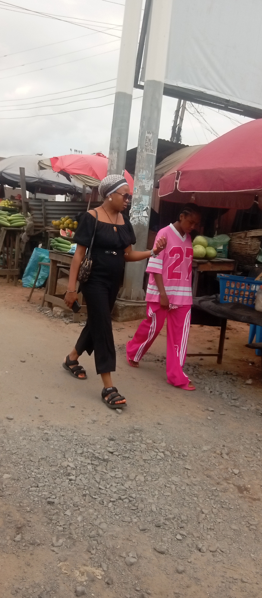 The image depicts a street scene in Onitsha, Nigeria, likely a market area. Two individuals are walking on a dirt and gravel road. In the foreground, a woman wearing a black off-the-shoulder top, black pants, and black sandals is walking to the left of the frame. She has on a patterned headwrap and sunglasses, and a dark shoulder bag. Her right hand is extended towards the person walking beside her. To her right, a younger person in a pink t-shirt with the number "27" on it and matching pink pants with white stripes is walking alongside her. Behind the individuals, market stalls with red and white awnings are visible, displaying various fruits and vegetables, including cucumbers and what appear to be gourds or melons. There are also crates and baskets, suggesting commercial activity. The overall environment appears dusty and somewhat unkempt, typical of a busy outdoor market. The sky is overcast.