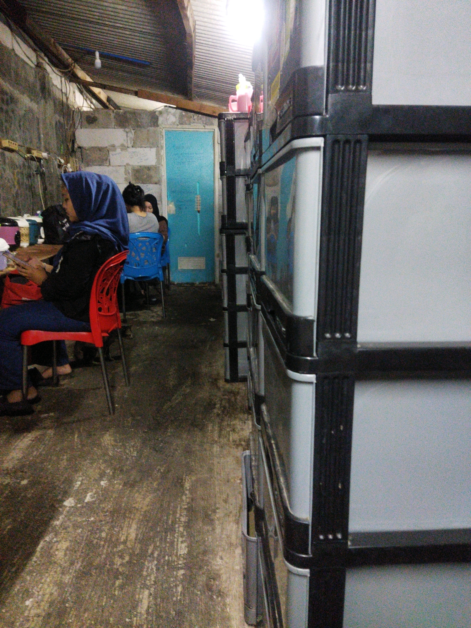 The image depicts an indoor scene within a building, likely a communal area or workshop, located in West Jakarta, Indonesia. In the foreground, a woman wearing a blue hijab and black clothing is seated on a red chair, holding a mobile phone and looking down. To her left, a portion of a black bag is visible. Behind her, a blue plastic chair is occupied by another individual. Further back, a blue door is visible, with a portion of a wall constructed of concrete blocks. To the right of the image, a tall, grey modular storage unit with black framing dominates the frame. The floor is concrete, appearing worn and somewhat dirty. The ceiling is corrugated metal, and a lightbulb hangs from the roof structure.