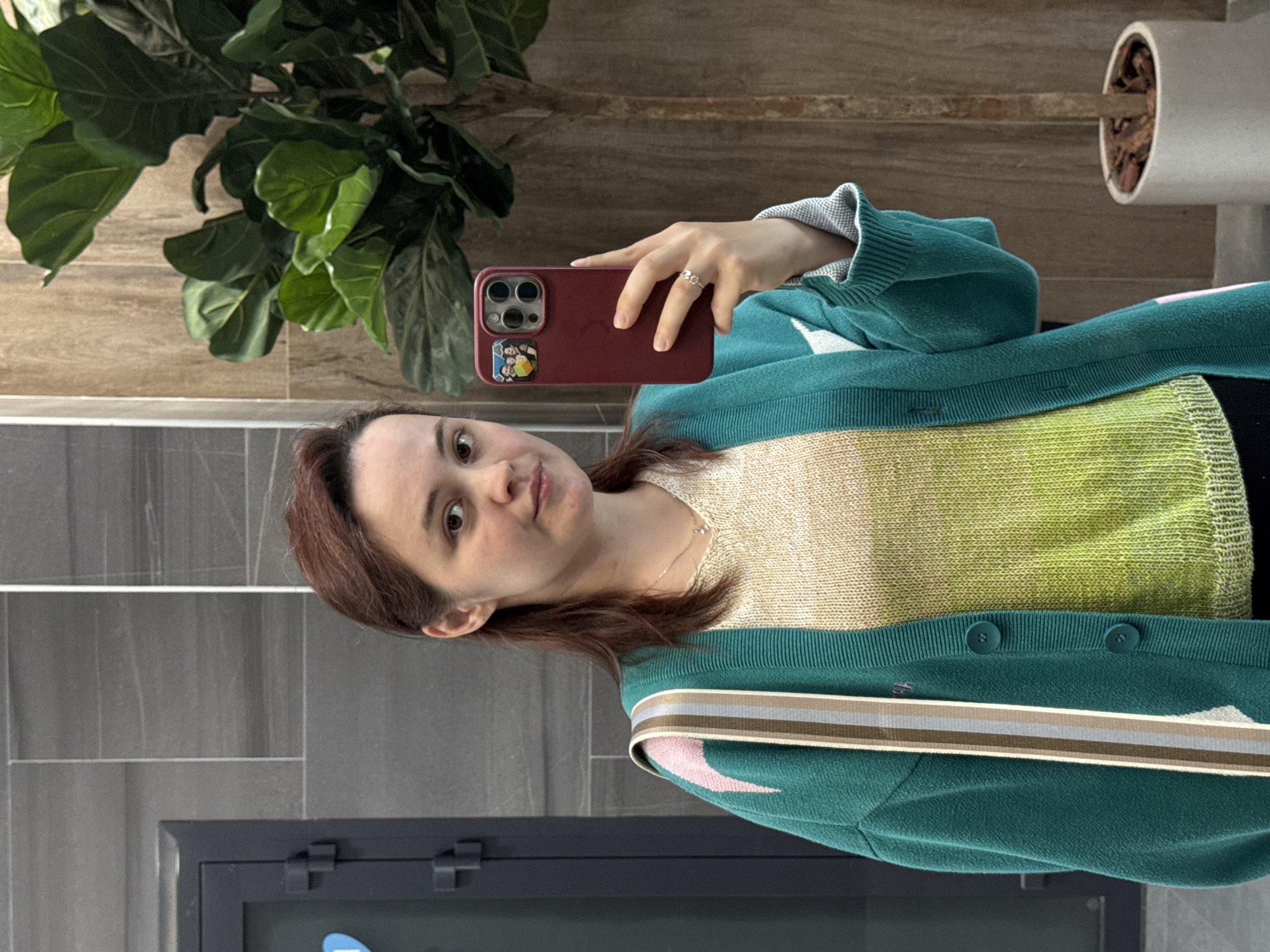 The image shows a young woman taking a mirror selfie. She has medium-length brown hair and is wearing a teal cardigan over a cream and green gradient knit sweater. A striped ribbon with pink, brown, and grey accents is visible on the cardigan. She is wearing a delicate necklace and a ring on her left hand. She is holding a dark red smartphone, which has a camera module and a small sticker on the back.
The background suggests an indoor setting. To the left, there is a large leafy plant, possibly a Fiddle Leaf Fig, with its leaves spread out. Behind the plant and the woman, there is a wall with a wooden texture. To the right of the plant, a white cylindrical pot containing some dry plant material is visible. Further behind, there are grey tiled walls and a dark cabinet or door with visible hinges.
The lighting appears to be artificial, creating a soft glow. The overall mood is casual and personal, typical of a mirror selfie. No specific time of day or weather conditions can be determined from the image. There is no visible text in the image. The context "Izhevsk, Russia" provides a geographical location but does not offer any visual cues within the image itself to confirm it.