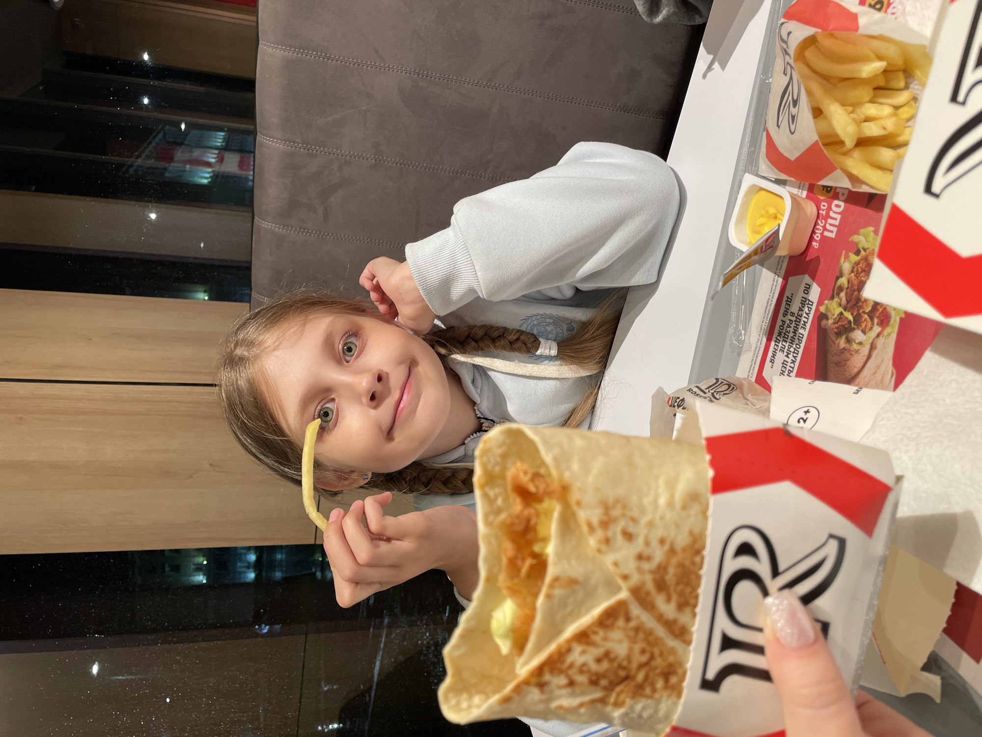 A young girl with braided blonde hair is sitting at a table in what appears to be a fast-food restaurant. She is wearing a light blue sweatshirt and has a French fry held up to one of her eyes. On the table are a package of French fries, a container of sauce, a promotional advertisement, and a partially unwrapped tortilla wrap from Burger King. A hand with polished fingernails is visible holding the wrap. The text in Russian on the promotional advertisement suggests it is related to promotions in the city of Izhevsk, Russia. It is dusk, and the table is near a window, which reflects the city lights. A gray upholstered bench seat is also visible.