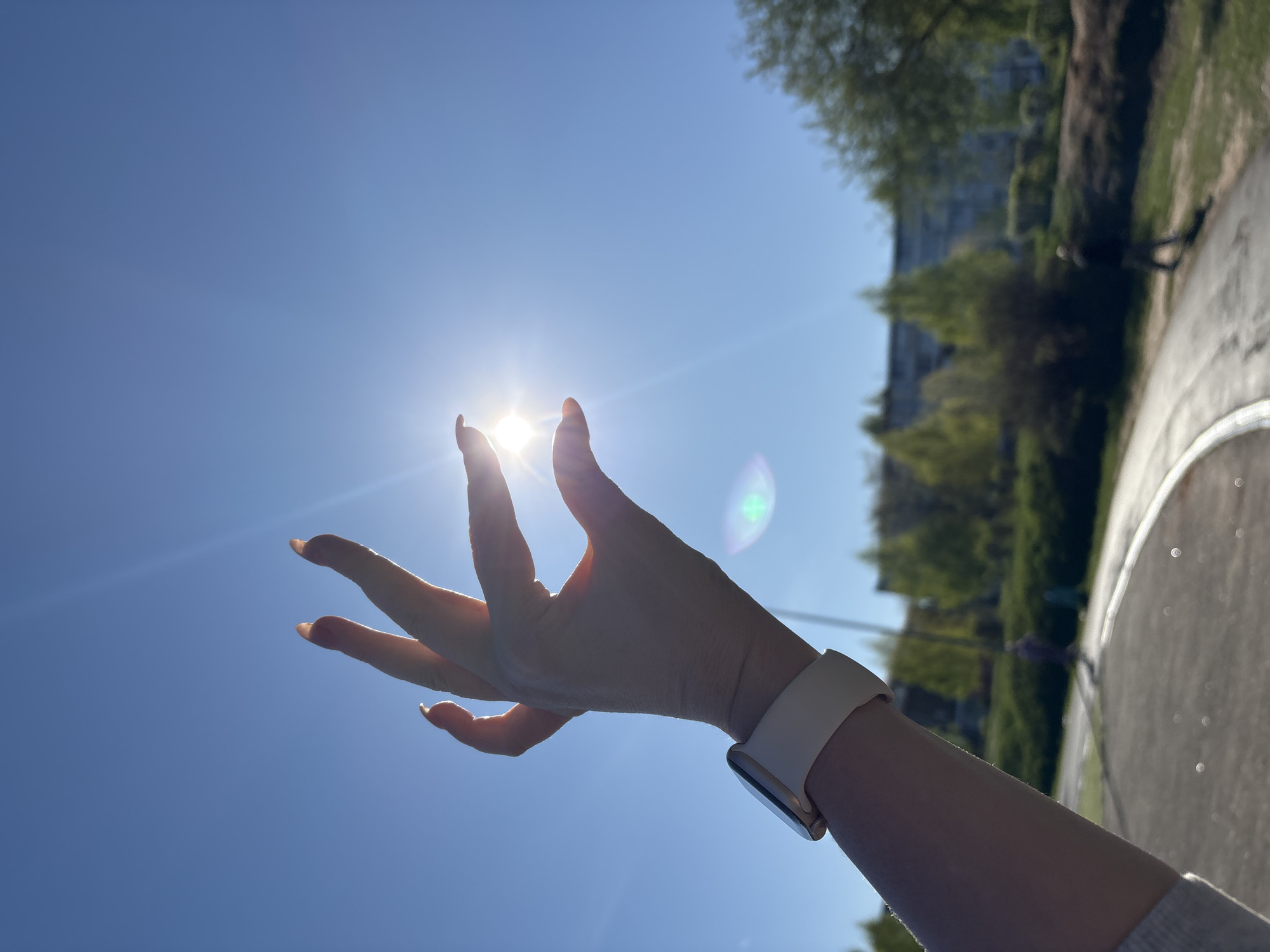 A person's hand is held up towards the sun in Chaikovsky, Russia, against a clear blue sky. The sun is positioned between the fingers, causing a bright flare. The person is wearing a white smartwatch on their wrist. In the background, trees and a paved walkway are visible, suggesting a park or public area. A building is also visible in the background.
