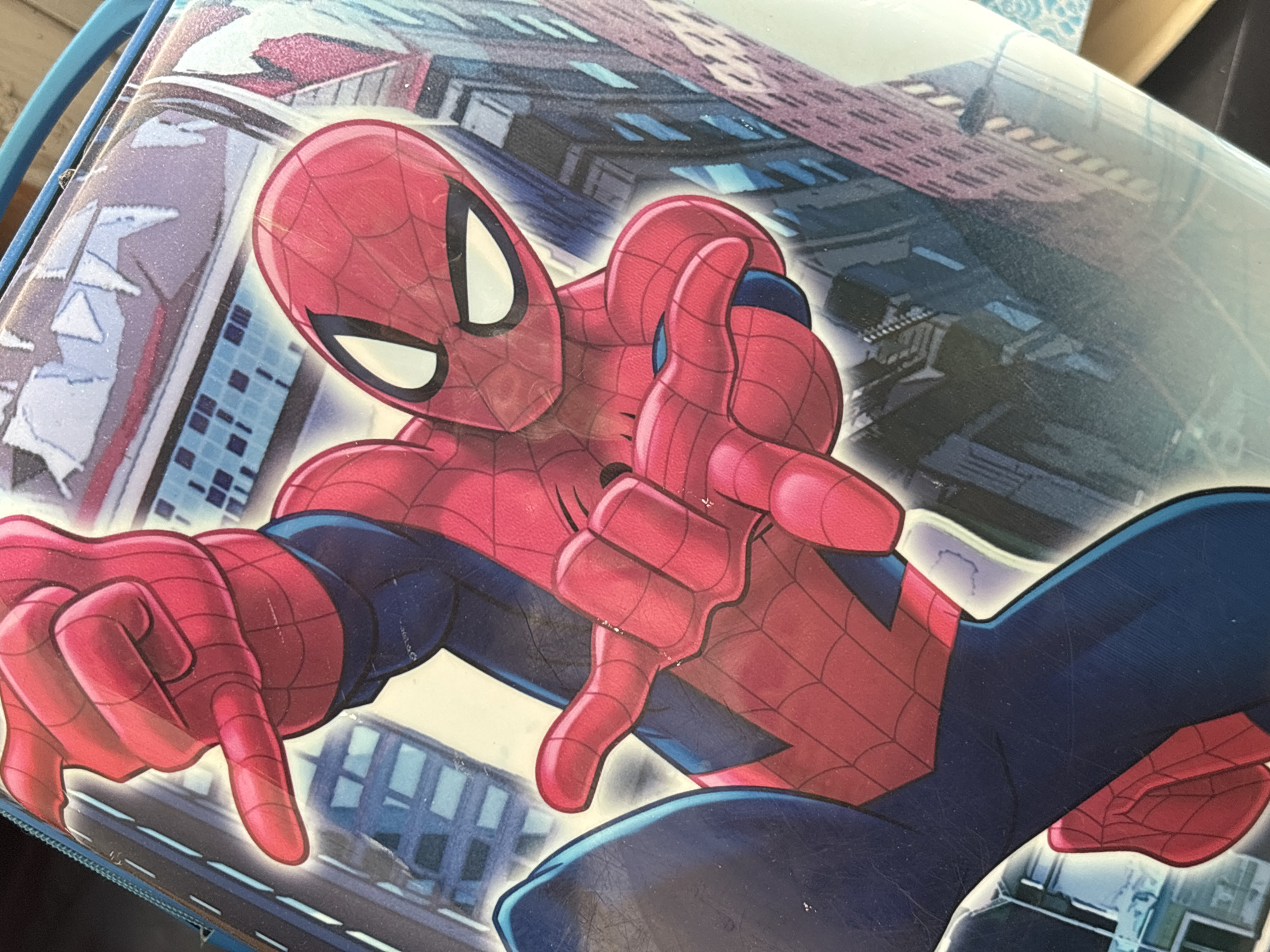 The image shows a close-up of a Spiderman lunchbox. Spiderman is depicted in a dynamic pose against a backdrop of city buildings. The buildings' architectural style is not distinct enough to determine a specific location such as Perm, Russia. The predominant colors are red, blue, and white. The Spiderman character is detailed with a web pattern on his red suit. The lunchbox appears to be made of plastic.