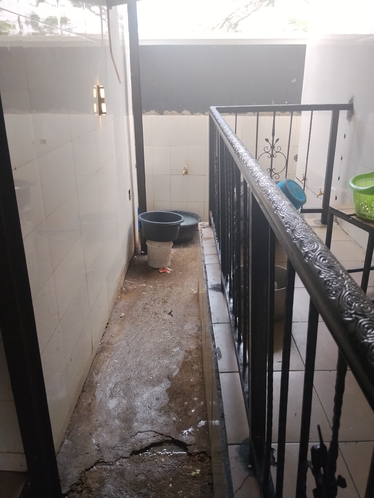The image depicts an outdoor, dimly lit, narrow walkway or service area. The walls are tiled with small, off-white square tiles, with visible grout lines. The floor is made of concrete and appears wet and dirty, with cracks and puddles of water.
In the middle ground, on the right side of the walkway, a black metal railing with decorative scrollwork runs along a slightly elevated area. Beneath the railing, on a concrete surface, are several large plastic buckets. One is a dark gray or black tub, resting on a white bucket. Another, lighter-colored bucket is visible. Behind these, a green plastic basket is seen on a metal shelf.
On the left side of the walkway, closer to the foreground, is a tiled wall. There are no people visible in the image. The scene appears to be an utilitarian area, possibly behind a building or in a courtyard. The presence of buckets and the wet floor suggest a place where water is used, perhaps for cleaning or some form of water storage.
The location is identified as Howayi, Nigeria. This specific scene does not provide overt visual cues to confirm its exact geographical context beyond what is stated. The environmental conditions suggest a potentially humid climate, given the wet surfaces.