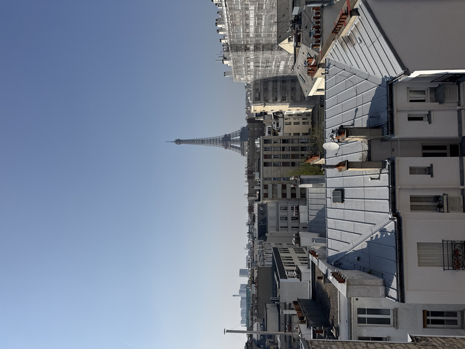 The image presents a daytime, elevated view of the Parisian cityscape, identified by the prominent Eiffel Tower centered in the middle distance. The sky is clear and light blue, with a faint contrail visible in the upper left quadrant.
In the foreground, a series of light gray, ribbed zinc rooftops are visible, characteristic of Parisian architecture. These roofs feature multiple dark brick chimneys, some topped with terracotta caps, and various vents or skylights. A white satellite dish is affixed to one of the central roofs. Below the roofs, the upper levels of apartment buildings are discernable, exhibiting white or light-colored facades, windows with closed white shutters, and a small balcony with a window box containing red flowers.
The midground is densely packed with multi-story buildings, primarily exhibiting light-colored stone or stucco facades. To the far right, a large building is entirely enveloped in white scaffolding and netting, indicating ongoing construction or renovation. To the left of the Eiffel Tower, on the distant horizon, a cluster of modern, taller buildings is visible, consistent with the La Défense business district. No individuals, actions, or direct interactions are visible within the scene. The lighting suggests direct sunlight, casting distinct shadows across the rooftops and building facades.