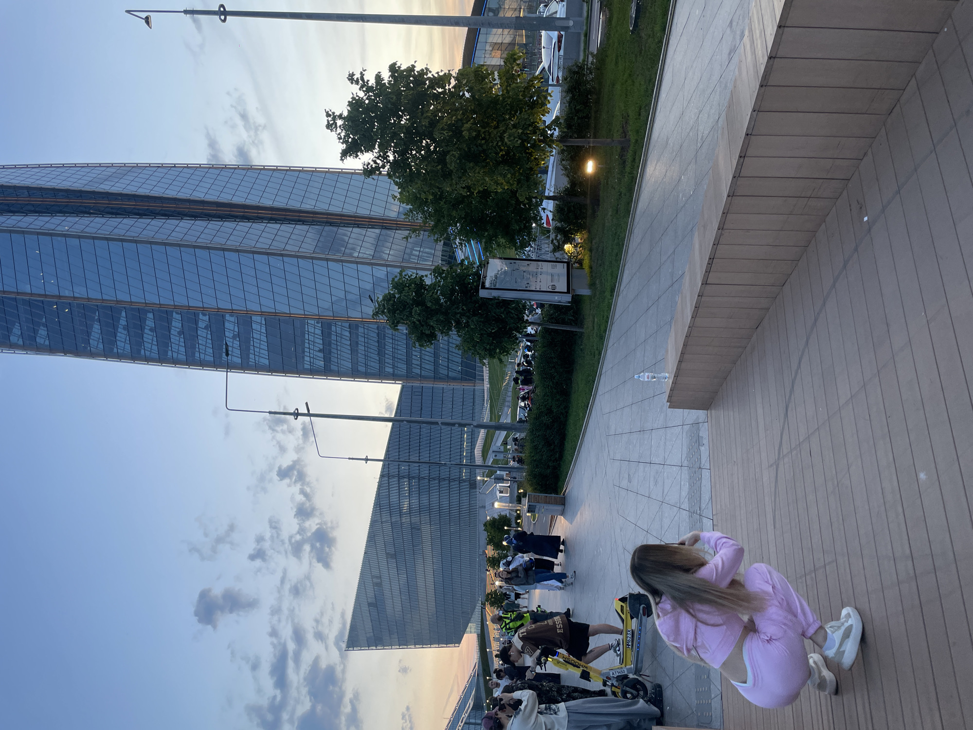 A group of people are gathered in a modern urban setting during the golden hour. In the background, a towering, sleek skyscraper with a glass facade dominates the skyline, reflecting the warm hues of the setting sun. To the left, a contemporary building with a geometrically angled roofline is visible. The foreground features a paved walkway with a wooden seating area. Several individuals are present, some walking, others standing. One person is seated on a yellow electric scooter. In the immediate foreground, a woman is crouched down, appearing to take a photograph with her phone. The atmosphere suggests a public space, possibly a park or plaza, with urban architecture. The sky is a mix of soft blues and oranges, with scattered clouds, indicating dusk.