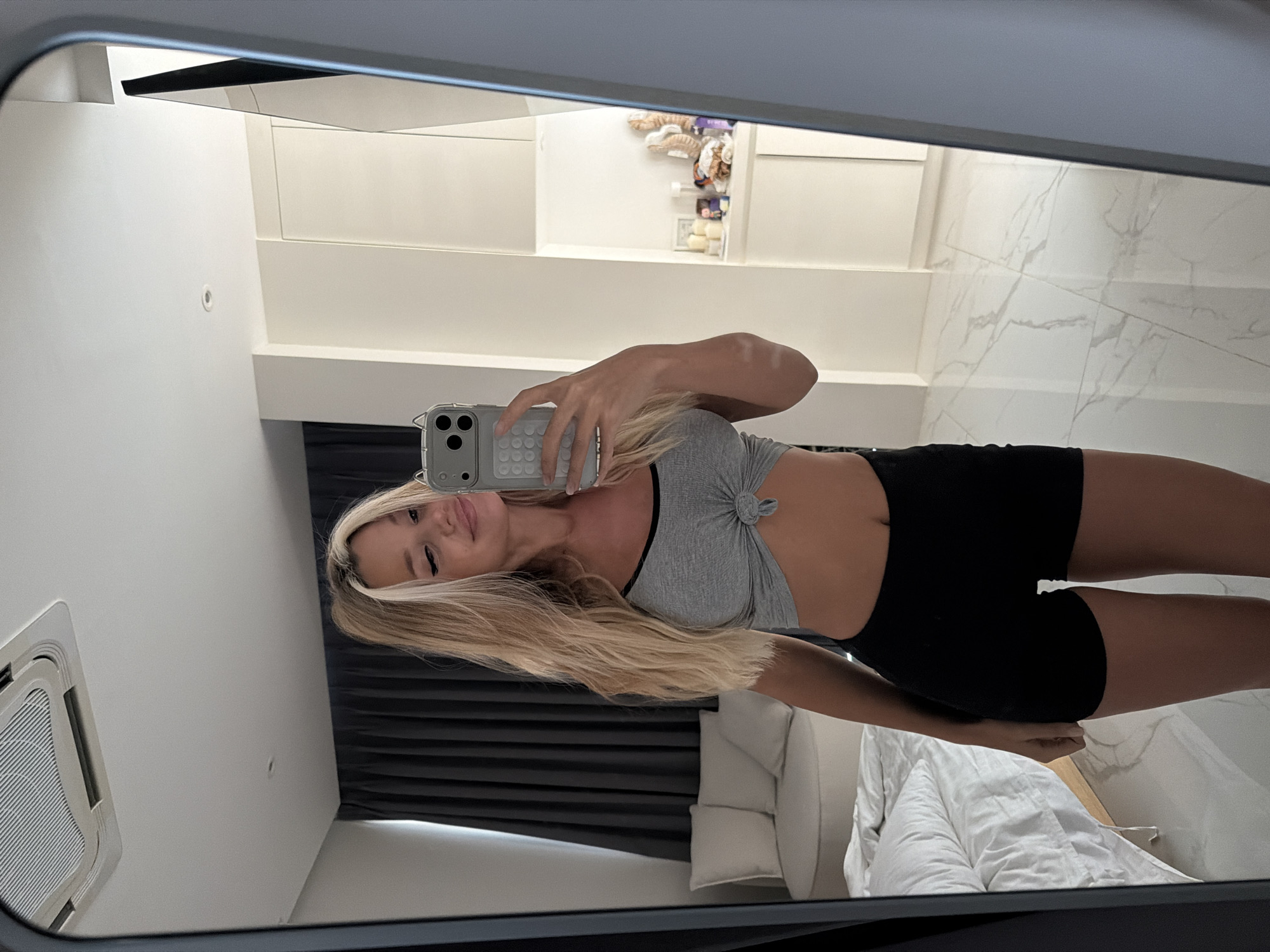 A woman with blonde hair is taking a mirror selfie in a bedroom. She is wearing a grey crop top and black shorts. The room is modern and minimalist, with white walls and marble flooring. A bed with white linens is visible in the background, along with a chair and dark curtains. The woman is holding a smartphone to take the photo. The location is Thep Krasatti, Thailand.