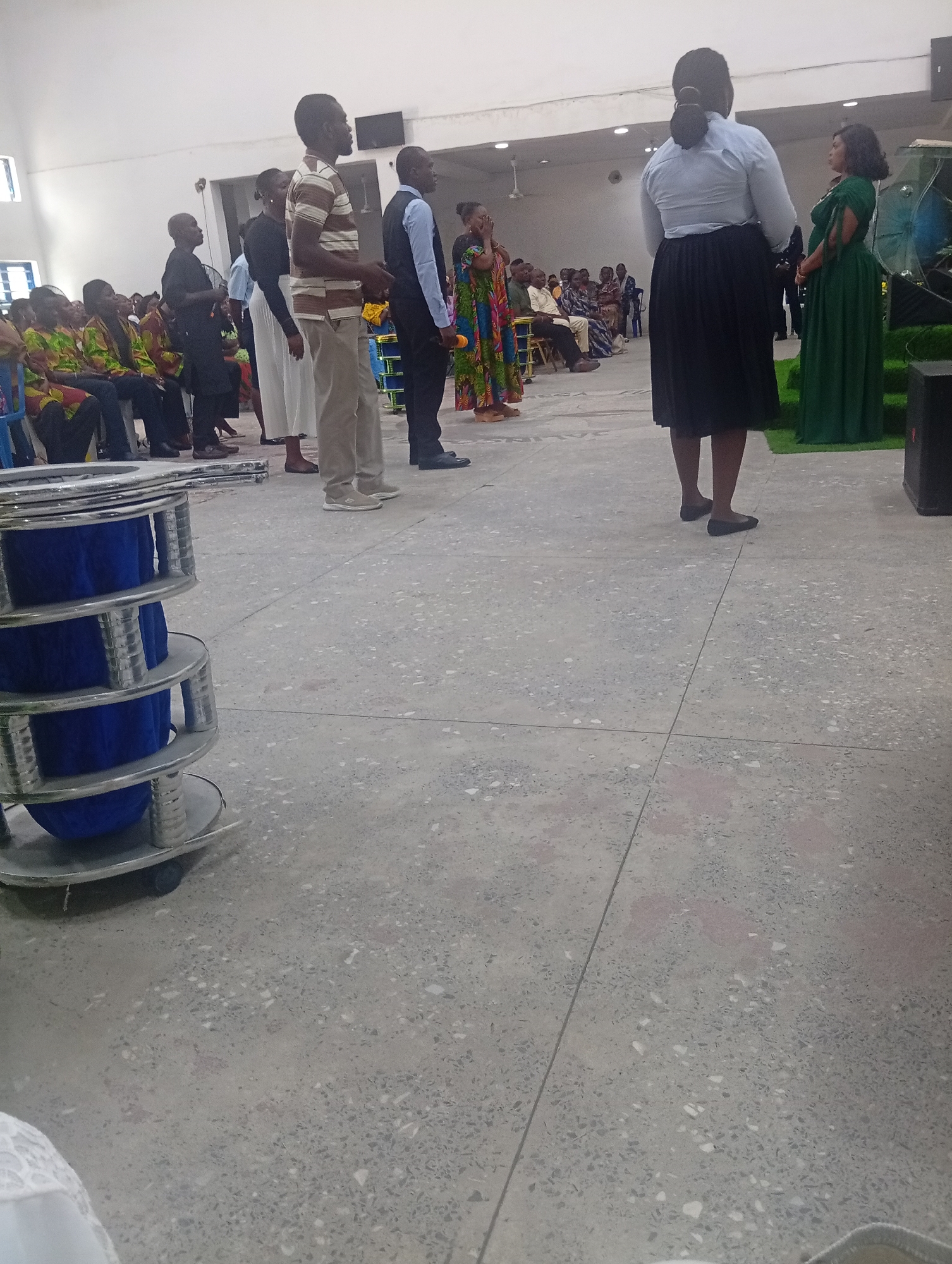 The image depicts an indoor gathering in Kason, Nigeria. A group of people are standing and seated in what appears to be a church or community hall. In the foreground, to the left, is a stacked object with silver-colored metal frames and blue fabric inserts, resembling tiered display stands or seating.
Several individuals are standing, facing away from the camera towards the right side of the image. One man in a striped shirt and khaki pants is prominent. To his right, another man in a blue shirt and black pants is visible. Further to the right, two women are standing; one in a light blue shirt and black skirt, and another in a long green dress. A woman in a colorful patterned dress stands to the far left, appearing to be observing or speaking.
In the background, to the left, a group of people are seated, some wearing brightly colored clothing, suggesting a cultural or religious event. The environment is characterized by a polished concrete floor and a white ceiling with lighting fixtures. The overall impression is of a formal assembly or service.