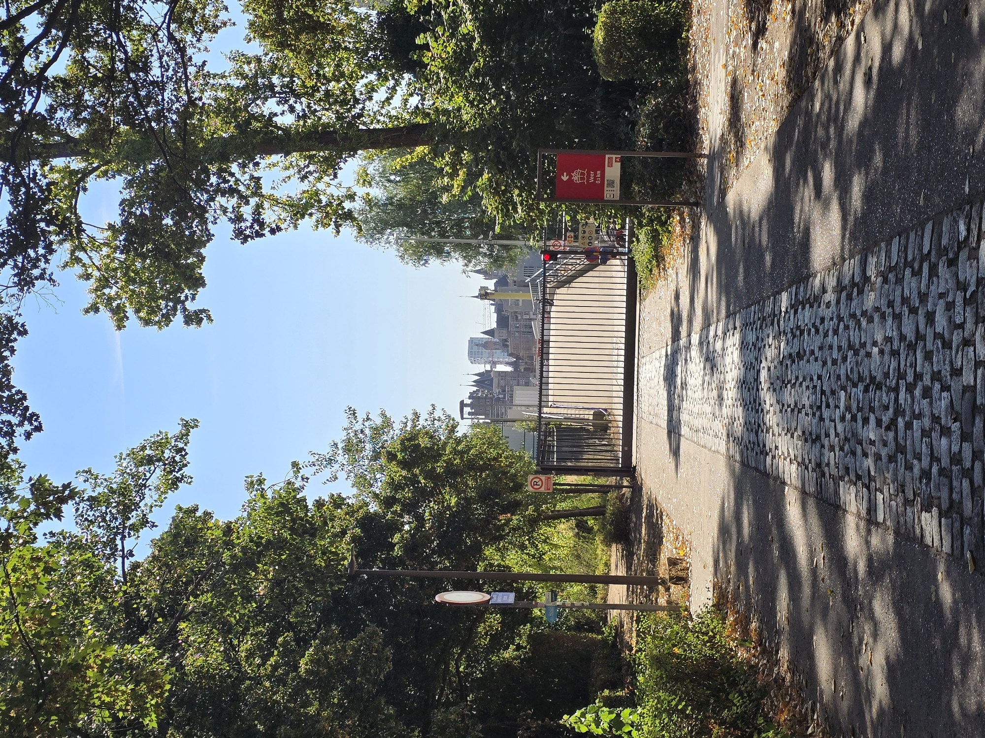 The image depicts a paved pathway leading to a large black metal gate. The pathway is a mixture of cobblestones and asphalt, with fallen leaves scattered on the ground. Lush green trees flank the pathway, casting shadows from the bright sunlight. In the background, a cityscape with distinctive spired buildings is visible beyond the gate, under a clear blue sky. A red directional sign with an arrow and the text "Veer 0,1 km" is positioned to the right of the gate. The scene appears to be an entrance to a park or a port area in Antwerp, Belgium.