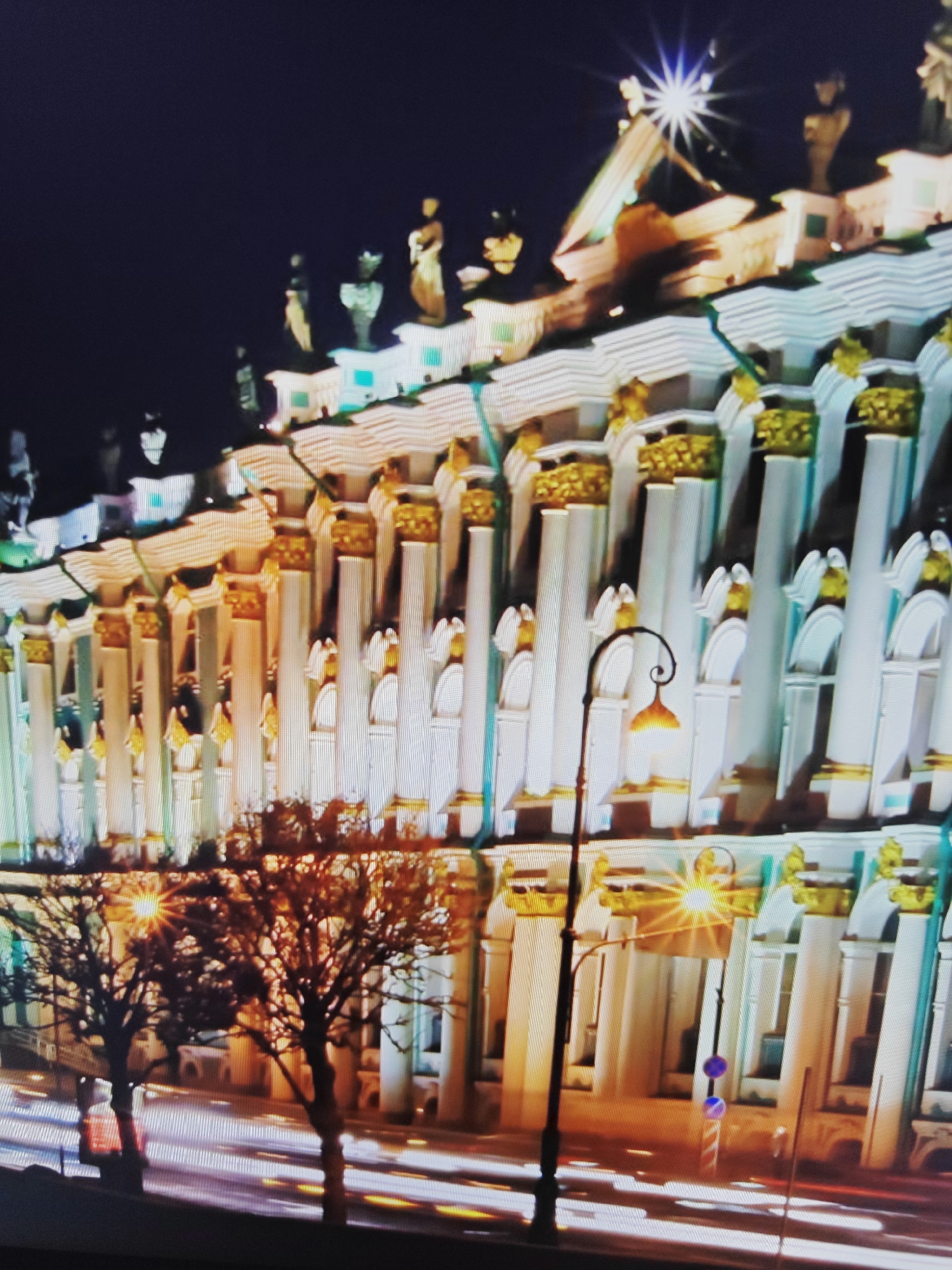 The image depicts a nighttime view of a grand building with classical architecture, characterized by numerous white columns topped with golden capitals. The building's facade is brightly lit, creating a dramatic effect against the dark sky. Statues are visible on the roofline. In the foreground, bare trees are silhouetted against the illuminated building, and streaks of light suggest moving vehicles on a street below. A street lamp with a decorative curve and a glowing yellow light is positioned to the left of the building's entrance. The overall scene suggests an urban environment at night. The location is identified as Unknown, Unknown.