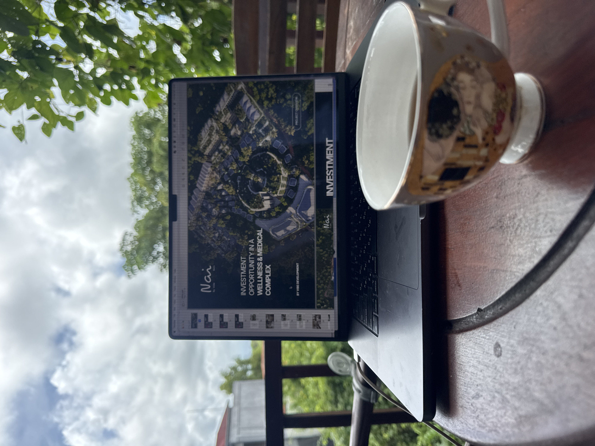 A laptop sits on a wooden table next to a coffee mug with a portrait design. The laptop screen displays an investment proposal for a wellness and medical complex, called "Nai by Vibe." Text on the screen indicates an investment opportunity. The image of the complex shows a circular design, with various buildings and greenery. The location of the photograph appears to be outdoors, possibly on a balcony, as greenery and sky are visible in the background. A metal railing is also visible. The laptop and mug are in sharp focus, while the background is slightly blurred. Kuta Selatan, Indonesia is not mentioned.