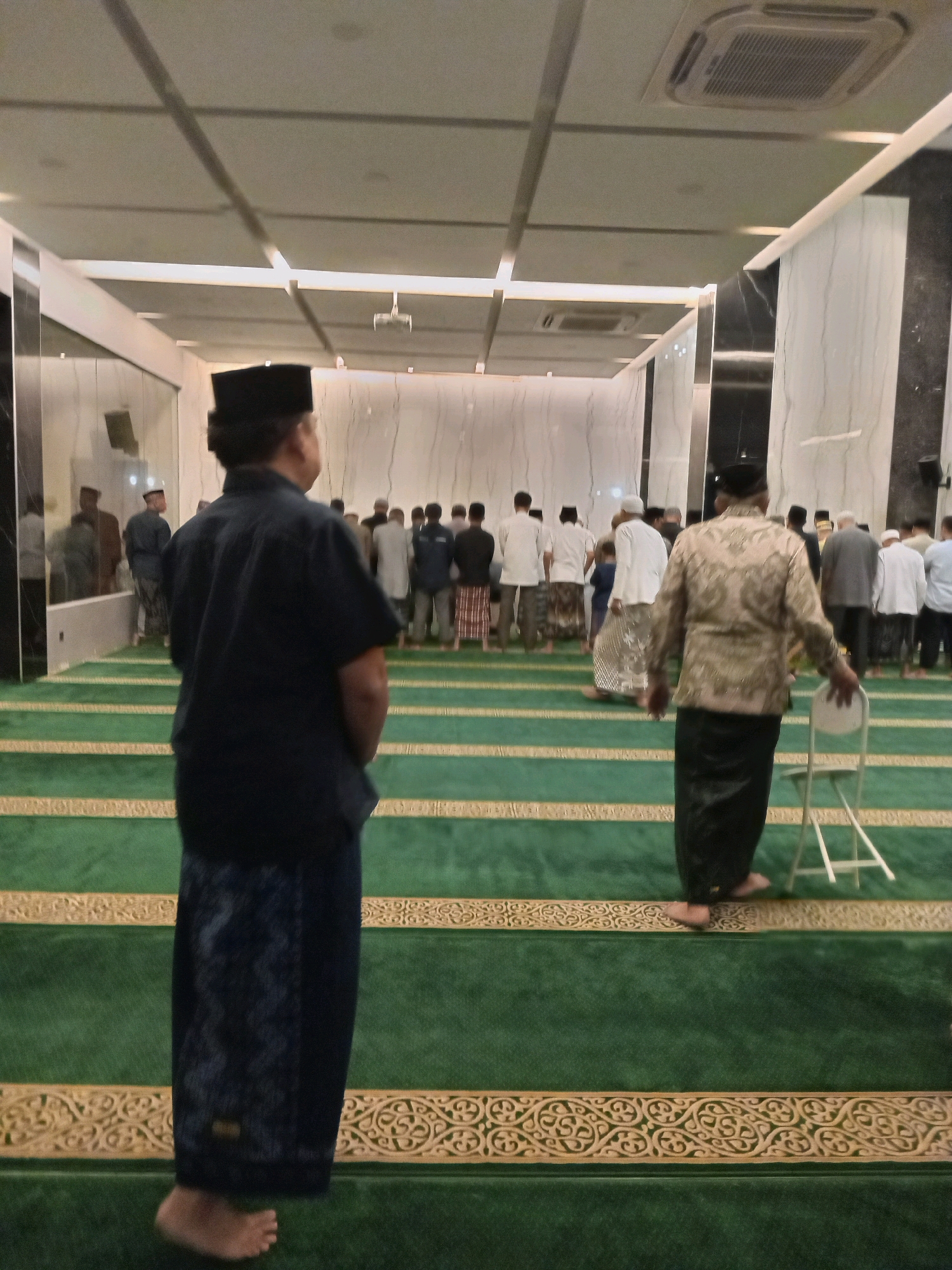 The image depicts a group of men in a mosque, with green carpeted floors marked with golden patterns. In the foreground, a man wearing a dark blue shirt and a patterned sarong stands facing away from the camera, his bare feet visible. To his right, another man in a patterned gold long-sleeved shirt and a black sarong also faces away, with a white folding chair nearby. In the background, more men are lined up, facing forward, dressed in various traditional Islamic attire, including sarongs and caps. The interior of the mosque features a modern ceiling with recessed lighting and air conditioning units. The overall scene suggests a religious gathering, likely a prayer service, in Randuagung, Indonesia.