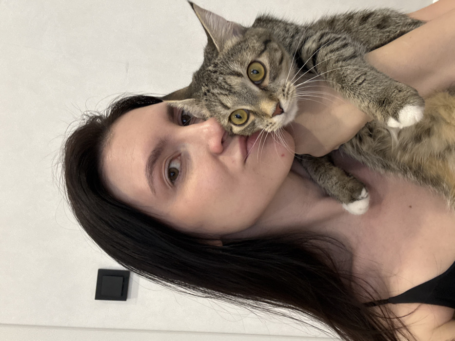 The image depicts an adult female holding a domestic cat in an indoor residential setting. No specific location within Kazan, Russia, is discernible from the visual evidence.
The female, positioned on the left, has long, dark brown hair, fair skin, and brown eyes. She is wearing a dark, possibly black, spaghetti-strap top. Her face is turned towards the right, with her lips pursed in a "kissing" gesture.
She is holding a short-haired tabby cat with striped and spotted fur in shades of brown and grey. The cat has prominent amber-yellow eyes and white paws. Its head is positioned slightly below the female's chin, looking directly forward. The female's left arm and hand are visible supporting the cat.
In the upper left background, a black square-shaped electrical switch or outlet is mounted on a plain, light-colored wall. The overall environment is minimalist and appears to be a well-lit interior room.