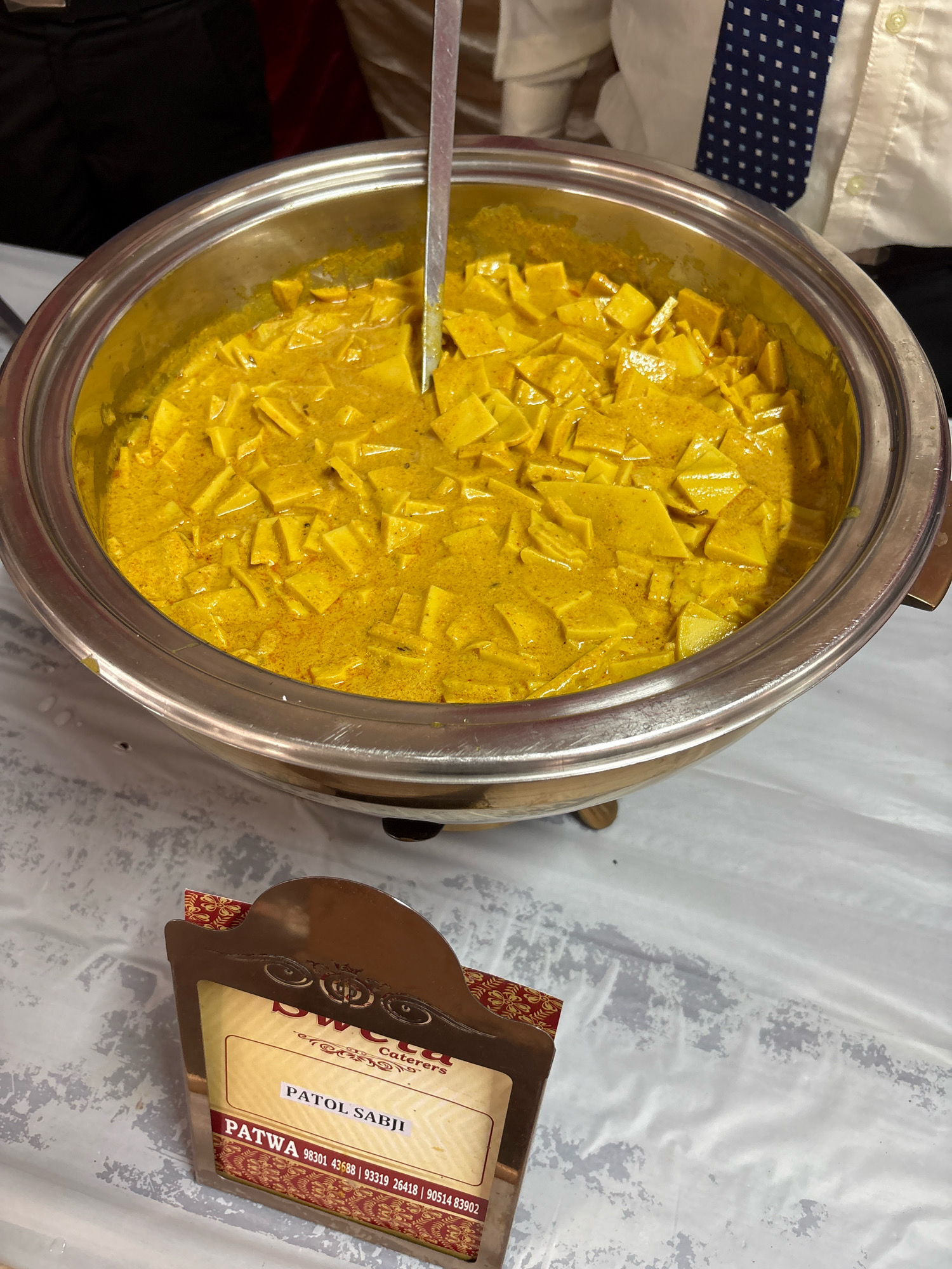 An overhead shot shows a large, circular, stainless steel serving dish filled with a yellow curry. The curry contains numerous diced pieces of a lighter yellow food item, possibly paneer or tofu. A metal ladle is partially submerged in the curry. In the foreground, a decorative cardboard stand displays a food label reading "PATOL SABJI" and "PATWA" with phone numbers. The serving dish is placed on a white tablecloth, and hints of people are visible in the background, including a person wearing a white shirt and a blue tie. The scene appears to be from an event or catering service, potentially in the city of Howrah, India, as indicated by the phone numbers on the label which correspond to Indian country codes.