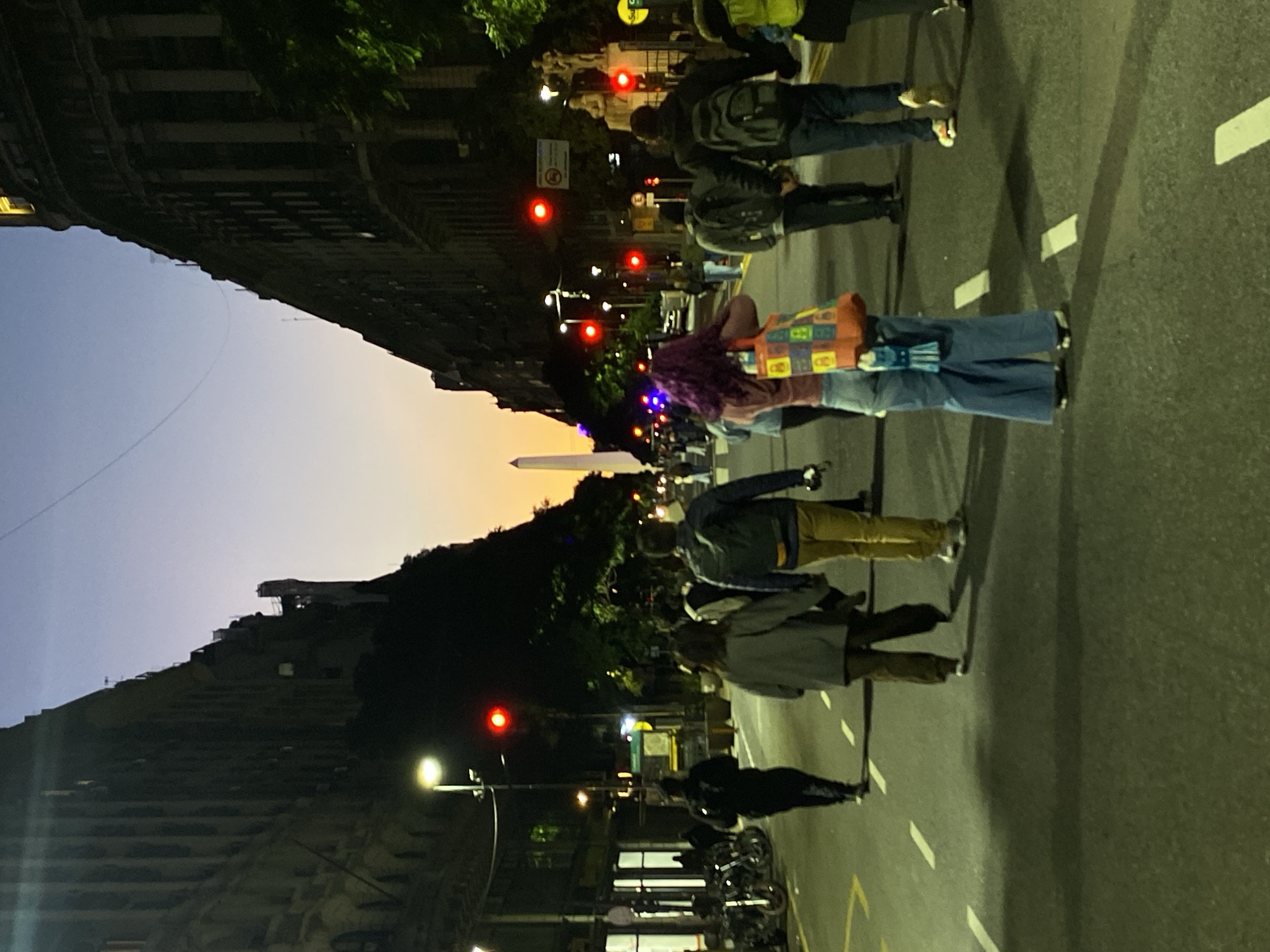 The image shows a street scene in Buenos Aires, Argentina at dusk. The Obelisco is visible in the distance. Several people are walking in the street, including a person with purple hair and a colorful patchwork bag. The street is marked with white lines and a yellow line. Traffic lights are visible, showing red. Buildings line both sides of the street. There are bicycles parked on the right side of the street.