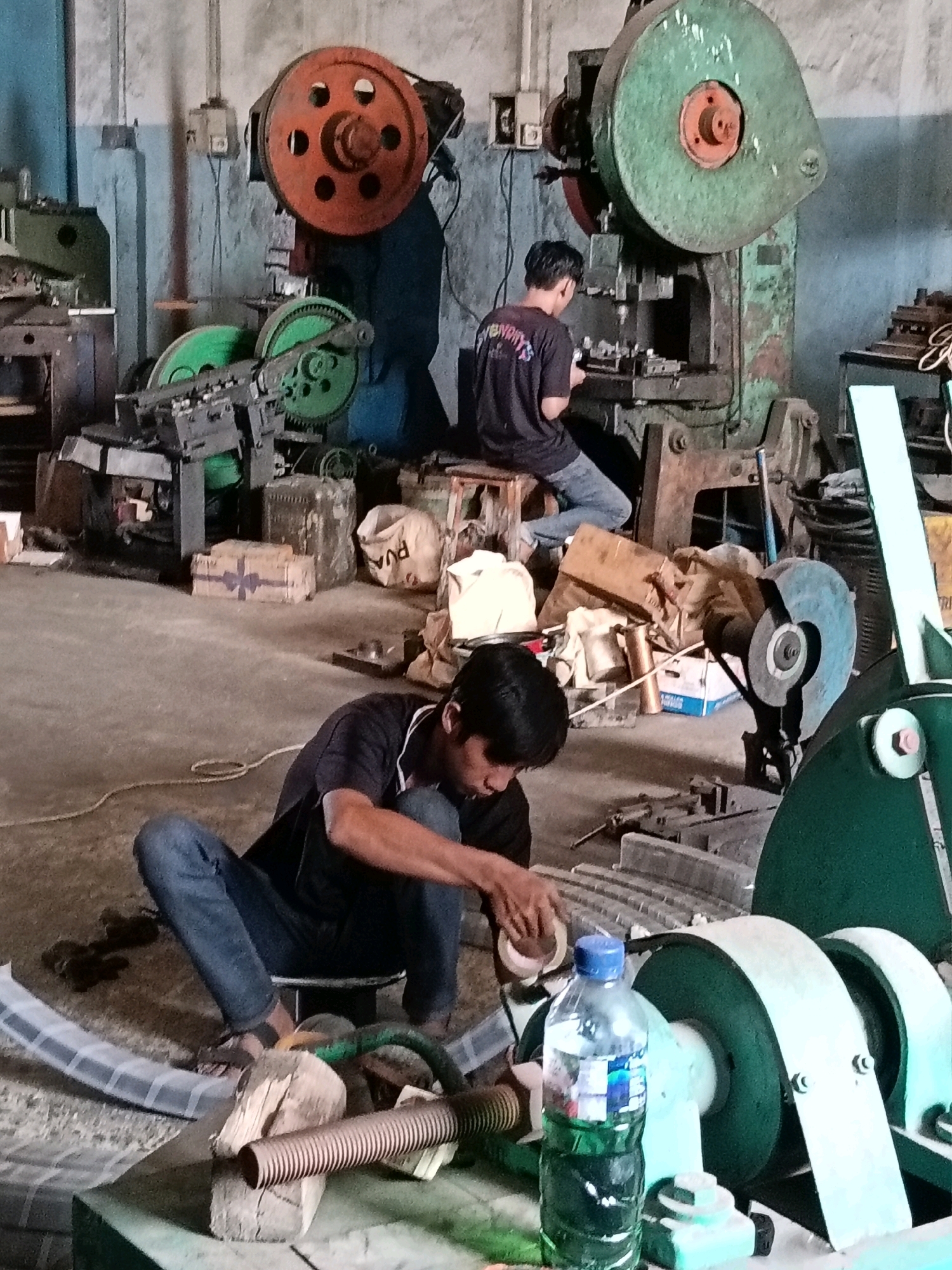 In a dimly lit workshop in Belimbing, Indonesia, two men are engaged in industrial work. One man, wearing a black t-shirt and blue jeans, sits on the floor in the foreground, focused on a piece of work with his hands. He is wearing sandals and has dark hair. A bottle of water with a blue cap is placed on a table next to him. In the background, another man, also wearing a black t-shirt and blue jeans, sits on a wooden stool in front of a large, green industrial press machine, seemingly operating it. The workshop is filled with heavy machinery, including large, colorful wheels on presses, gears, and metal components. Various objects, such as cardboard boxes, sacks, and electrical conduits, are scattered around the floor, suggesting an active and working environment. The walls are concrete, and overhead electrical boxes are visible.