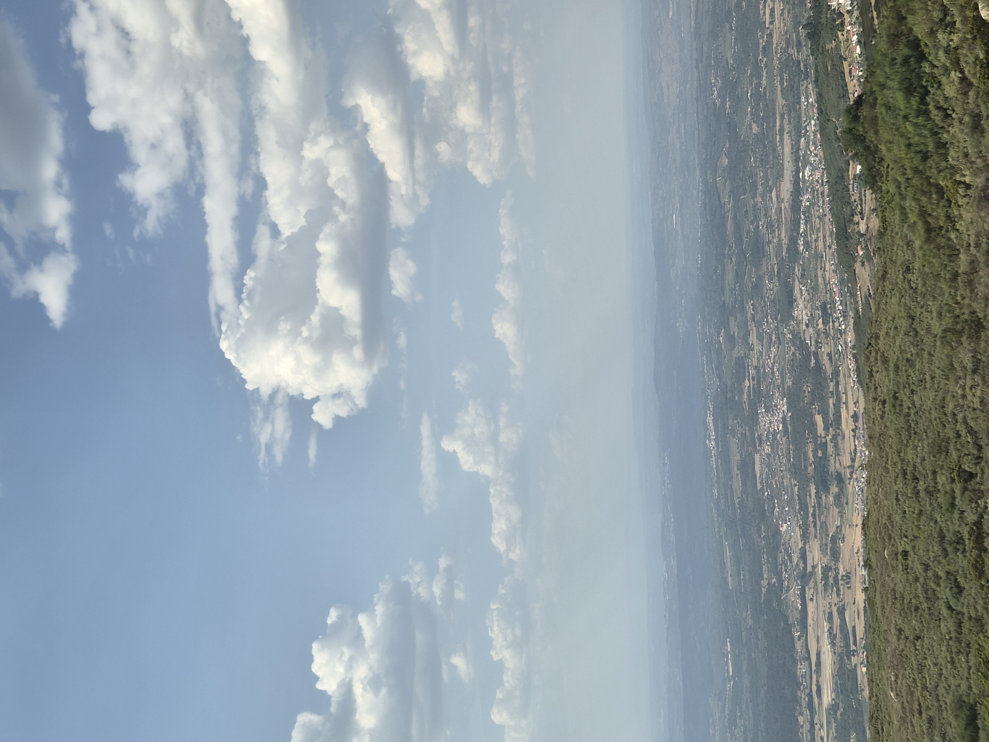 This is a wide-angle, high-angle landscape photograph taken from a vantage point above the city of Seia, Portugal. The image captures a vast expanse of rolling hills and a valley dotted with buildings, appearing to be a town or village. The foreground is dominated by dense, green vegetation, likely shrubs or low-lying trees, covering the hillside.
The middle ground showcases the human settlement, a collection of houses and buildings with red-tiled roofs spread across the landscape. Agricultural fields, some appearing fallow or with harvested crops, are interspersed with patches of darker green vegetation. Roads or pathways are visible, winding through the town and connecting it to the surrounding areas.
The background consists of distant, hazy hills that fade into the horizon. The sky is a pale blue, filled with a mix of large, fluffy white cumulus clouds and smaller, more dispersed clouds. The lighting suggests it is daytime, with the sun likely overhead, casting a soft light on the scene. There are no people or specific actions depicted in the image. The overall impression is one of a serene, rural landscape.