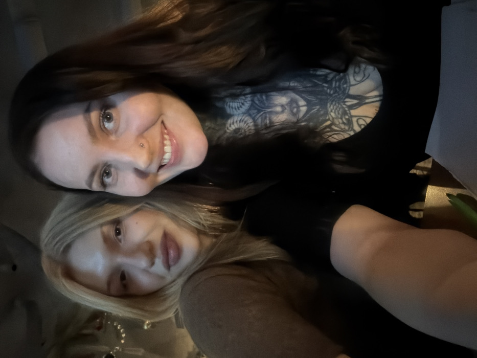 Two women are shown in a close-up, angled shot, likely a selfie taken indoors in what appears to be a dimly lit setting, possibly an evening or night out. The image is oriented with the women's heads tilted to the right.
The woman in the upper part of the frame has long, dark brown hair, fair skin, and light-colored eyes. She is smiling broadly, showing her teeth, and has a small piercing on the side of her nose. She is wearing a dark-colored top, possibly black. A prominent black and white tattoo covers a significant portion of her chest and shoulder area, depicting what looks like a mythological figure, possibly Medusa, with snakes forming part of the design. Her expression conveys happiness.
The second woman is positioned below the first, with her head resting slightly on the first woman's shoulder or chest. She has blonde hair that falls around her face and light-colored eyes. Her expression is more subtle, a gentle smile or neutral, content look. She is wearing a light brown or tan-colored top with long sleeves.
The background is dark and out of focus, suggesting an indoor environment with subdued lighting. There are some faint reflections or blurry shapes, possibly indicating decorative elements or distant lights. No specific objects or text are clearly discernible in the background. The overall mood of the image is warm and friendly.