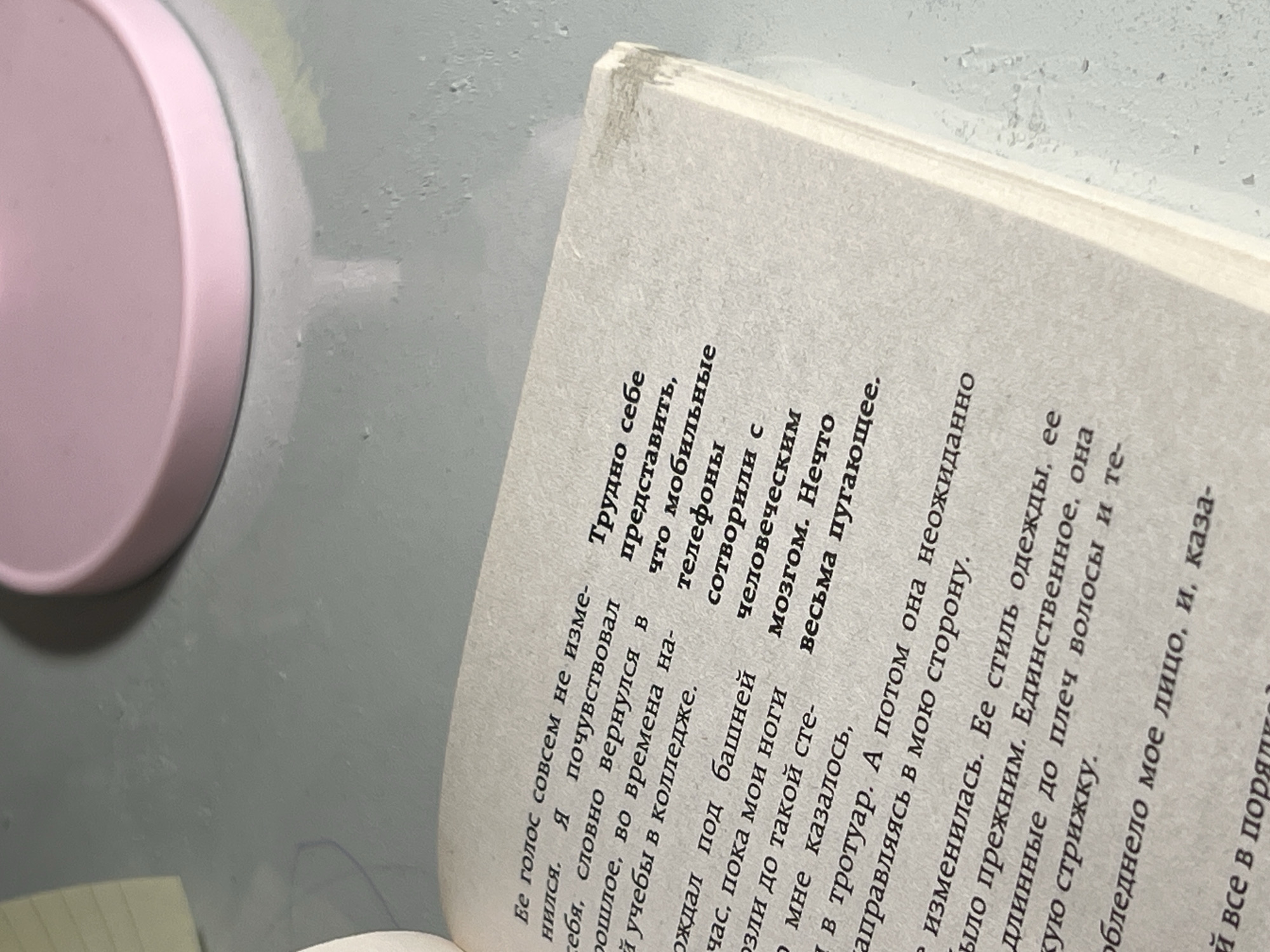 The image shows an open book with Russian text, placed on a light grey surface. A portion of a pink object, possibly a lid or a container, is visible on the left side of the frame. The book is angled, with the text running diagonally. The pages appear to be aged, with some discoloration and wear along the spine. The text discusses a voice, memories of college, and changes in a person's appearance, including their clothes and hairstyle. The tone of the text seems introspective and somewhat unsettling, referring to the frightening effect of mobile phones on the human brain. The lighting is soft, suggesting an indoor environment, though the time of day is not discernible. The overall scene is a close-up view of a book, highlighting the textual content and its physical condition.