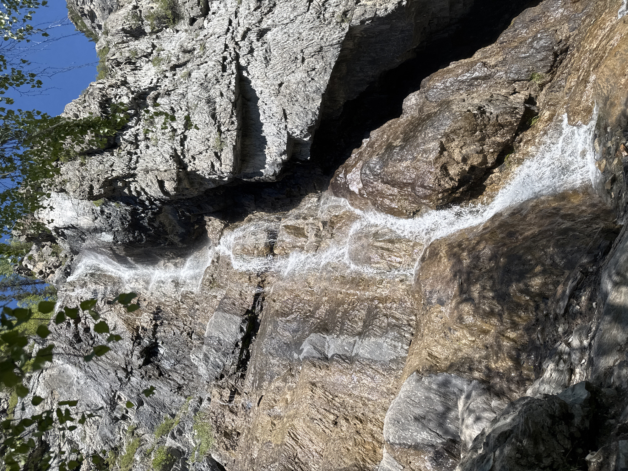 The image depicts a waterfall cascading down a rocky cliff face. The rock is varied in color, ranging from light gray to darker brown hues, and is textured with sharp ridges and crevices. Water streams down the rock face, creating a white, flowing effect. Some greenery, likely foliage from trees, is visible in the lower left corner. The sky, partially visible in the upper left corner, is a clear blue. The scene appears to be located in a natural, possibly mountainous environment.