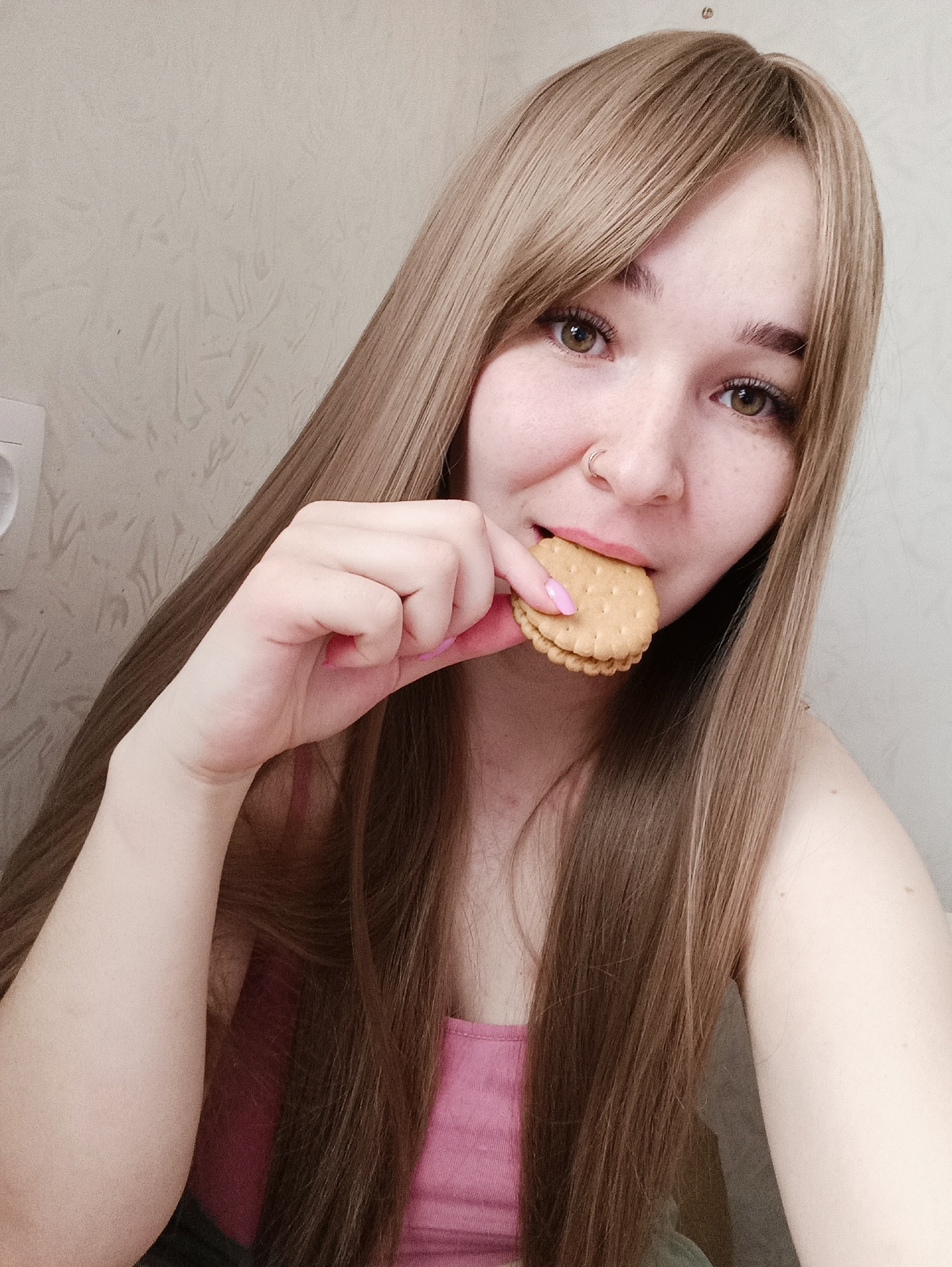 The image depicts a close-up portrait of a young adult female from the chest up, engaged in eating. The subject has long, light brown hair with bangs, fair skin, and green eyes. A small, circular metallic nose ring is visible in her left nostril. She is wearing a pink tank top. Her right hand, with fingernails painted pink, holds a round sandwich cookie to her mouth. The cookie consists of two light-brown, textured biscuits with a lighter-colored filling. The background is a plain, light-colored wall, possibly textured, with a portion of a white electrical outlet visible on the left side of the frame. The specific location within the city of Izhevsk, Russia, cannot be determined from the visual information provided.