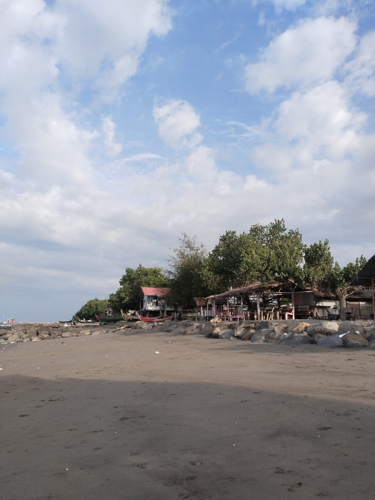 The image depicts a beach scene in Lhokseumawe, Indonesia. The foreground is dominated by dark, damp sand with faint tire tracks and scattered debris. In the middle ground, a rocky embankment lines the shore, behind which several structures are visible. To the left, a red-roofed building with white walls sits among lush green trees. Several boats, including a red one and a green one, are pulled up onto the sand near the building. Further to the right, a covered open-air structure with a thatched roof is supported by wooden posts. This structure appears to be a seating area, with tables and chairs visible underneath. More trees and a glimpse of another thatched-roof building are present to the far right. The background features a calm sea and a pier extending into the water on the far left, with a distant structure on the pier. The sky above is a mix of blue with scattered white clouds, suggesting fair weather. The overall impression is one of a quiet, possibly somewhat rustic, coastal settlement.