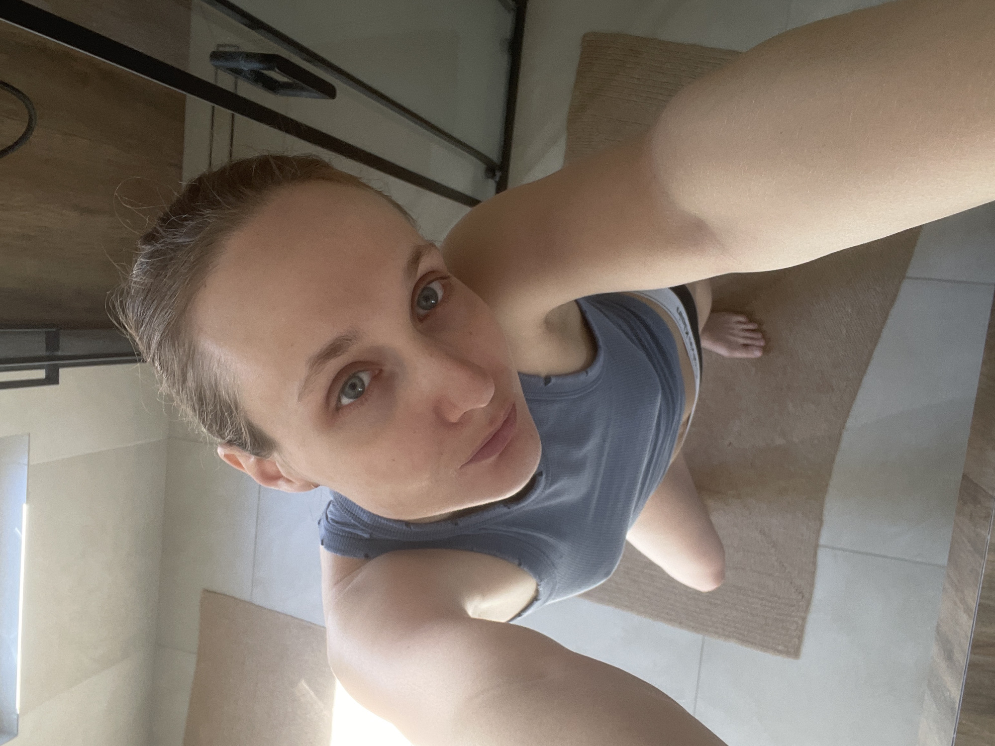 The image is a selfie taken by a woman with short brown hair and light eyes. She is wearing a gray tank top and appears to be in a bathroom, as evidenced by the tiled floor and a rug. A bed frame is partially visible in the background. The exact location within Подшивалово, Russia, cannot be determined from the image.