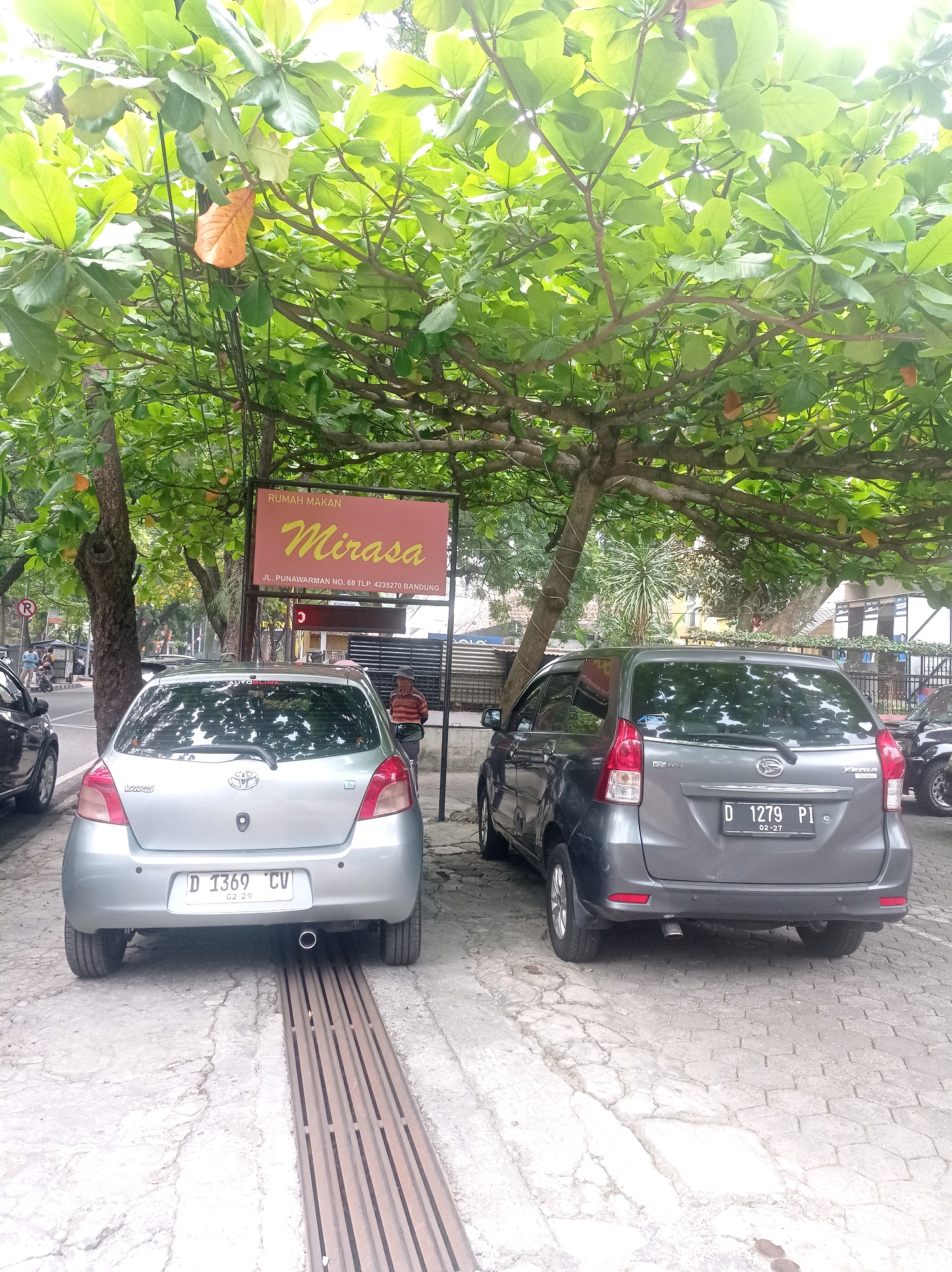 The image shows a street scene in Bandung City, Indonesia, beneath a large tree with green foliage and some falling orange leaves. A sign for "RUMAH MAKAN Mirasa" is visible, along with the address "JL. PUNAWARMAN NO. 68 TLP. 4235270 BANDUNG". Two cars are parked on the side of the street. The car on the left is a silver Toyota Yaris with license plate "D 1369 CV". The car on the right is a dark gray Daihatsu Xenia with license plate "D 1279 PI". A man in a striped shirt is standing near the Xenia. The ground is paved with interlocking concrete blocks and has a metal grate drain. In the background, there are more trees, buildings, and a few people walking on the sidewalk.