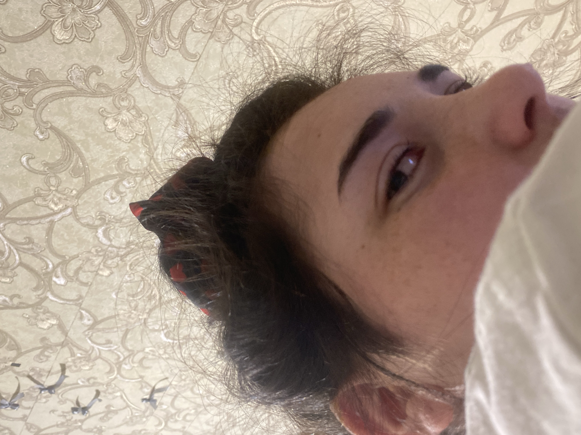 An indoor close-up shot of a person's face from a low angle. The person has dark hair with a hair tie, fair skin with visible freckles, and dark eyebrows. One eye is clearly visible and brown. A portion of a white garment is also visible. The background is a patterned wallpaper with floral designs. There's no definitive information to place this photo specifically in Poltava, Ukraine.