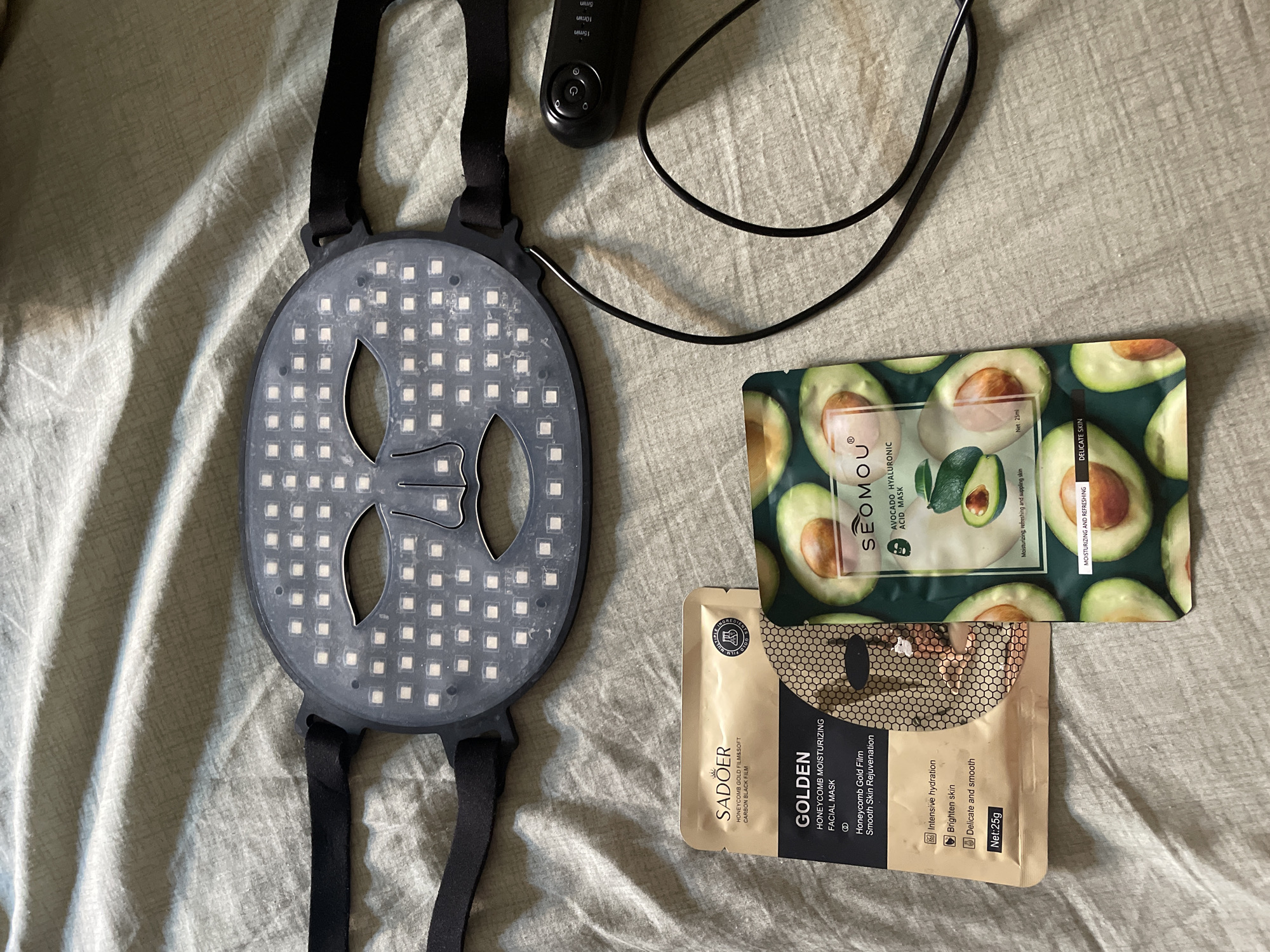 The image displays a collection of skincare products and a facial treatment device laid out on a textured, light brown fabric surface. The primary object is a black LED facial mask with numerous small, square white lights arranged across its surface. The mask is contoured to fit a face, with openings for the eyes and mouth. Two adjustable black straps are attached to the sides of the mask. A black wire extends from the mask to a small black remote control, which has several buttons and appears to be displaying timer options such as "15min", "10min", and "5min".
To the left of the mask are two foil packets of facial masks. The packet on the left is gold and features text that includes "SADOER", "GOLDEN HONEYCOMB MOISTURIZING FACIAL MASK", and "Honeycomb Gold Film Smooth Skin Rejuvenation". It also lists features like "Intensive hydration", "Brighten skin", and "Delicate and smooth", along with "Net: 25g". The packet on the right is dark green and features images of halved avocados. Its text includes "SEOMOU", "AVOCADO HYALURONIC ACID MASK", and "Moisturizing, refreshing and supple skin". Smaller text at the bottom reads "DELICATE SKIN".
The overall impression is a static arrangement of beauty and skincare items. The image is a top-down, slightly angled view of these products. There are no people or animals present in the image, nor any indicators of a specific city, town, or country, including Kadıköy, Turkey. The environment is a plain, textured fabric.