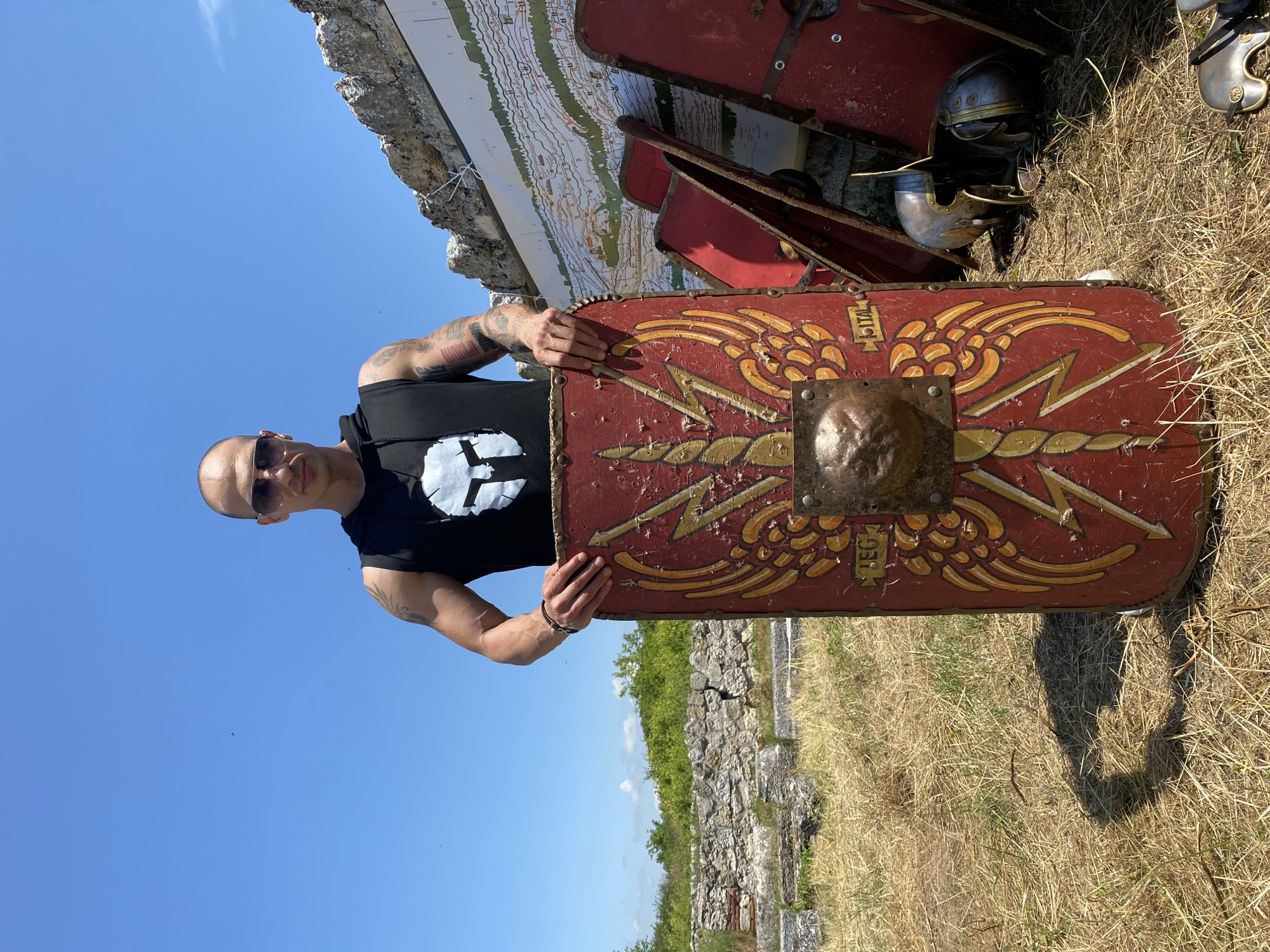 A male individual, with a bald head and wearing sunglasses, stands outdoors under a clear blue sky. He is wearing a black sleeveless hoodie featuring a white stylized Spartan helmet graphic on the front. Tattoos are visible on both his arms. He is centrally positioned, holding a large, curved red Roman scutum shield vertically in front of him with both hands.
The shield is decorated with prominent yellow/gold designs, including two winged lightning bolts originating from a central circular metal boss that depicts a stylized lion's head in relief. The text "LEG" is visible on the lower left of the shield and "ITAL" on the lower right, indicating Legio I Italica. To the immediate right, several similar red shields are stacked, and two Roman-style helmets lie on the dry, yellowish-brown grassy ground.
In the background, to the left, are stone ruins consistent with an ancient archaeological site, with green foliage and distant trees under a bright blue sky. To the right, behind the figure, a large, detailed informational map or diagram displaying topographical features and possibly ancient routes is affixed to a stone wall or structure. The overall scene suggests an archaeological site, specifically identified as the Roman city of Oescus near Gigen, Bulgaria, given the specific legionary inscription on the shield. The sun is high, casting distinct shadows on the ground.