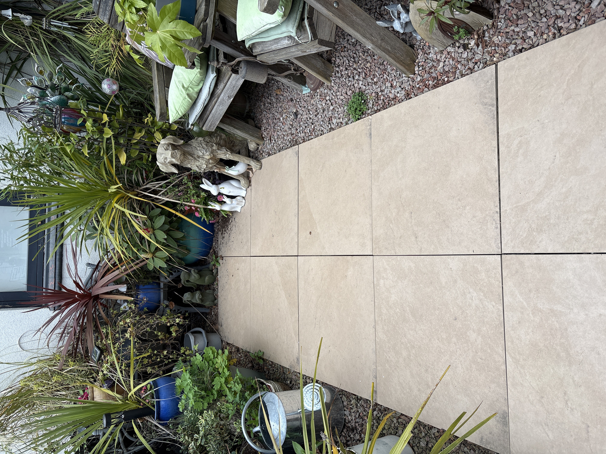 The image shows a garden path in Shrewsbury, United Kingdom, constructed of large, light-beige square paving slabs laid in a grid pattern. The path is flanked on one side by a densely planted garden area filled with various potted plants, including a cordyline plant with maroon leaves, various palms and other leafy plants, and a collection of garden ornaments. Among the ornaments are ceramic rabbits, a decorative dog statue, and several small elephant figures. A galvanized watering can sits near the edge of the path, partially obscured by foliage. Adjoining the paving slabs on the other side is a small area covered with reddish-brown gravel where a wooden chair is located with green cushions placed on it.