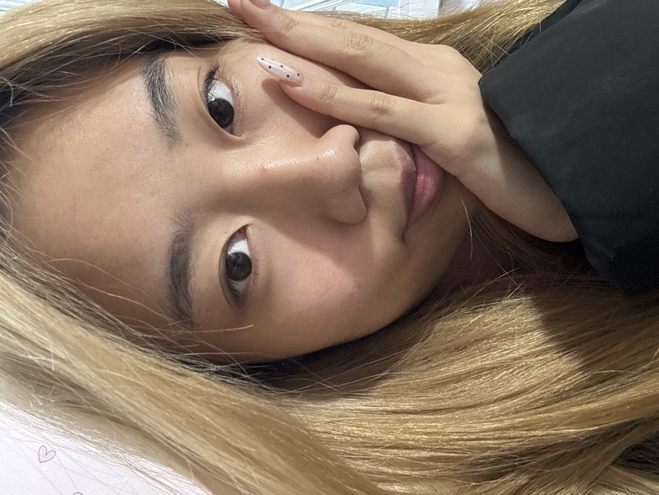 The image depicts a close-up, head-and-shoulders portrait of a young East Asian female, facing forward and looking directly at the camera. She has long, blonde hair that frames the left side of her face and extends over her shoulder. Her skin tone is fair, and her eyes are dark brown. She has dark eyebrows and appears to be wearing minimal or no makeup. Her left hand is positioned with the palm against her left cheek and chin, partially obscuring the lower-left portion of her face. Her fingernails are manicured, with a light pink or nude base polish adorned with small red polka dots. A black sleeve, likely from a shirt or jacket, is visible on her lower-right, covering her forearm. The background is softly out of focus, appearing to be an indoor setting with light-colored walls, possibly white or light pink, featuring faint, small, pink heart-shaped patterns on the upper left. The specific location within Shapingba District, China, cannot be determined from the visual information provided in the image; the scene is consistent with an indoor environment.