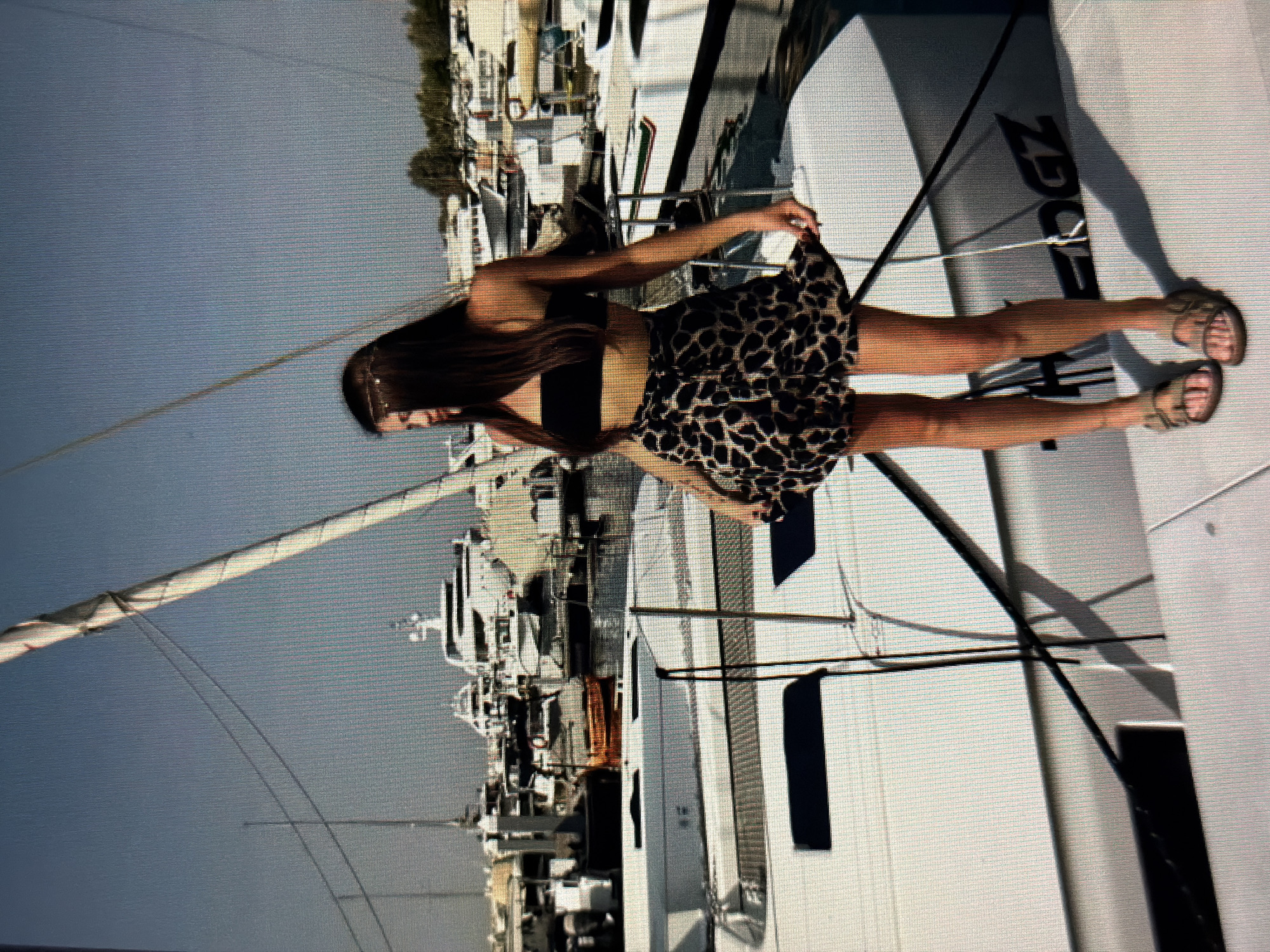 A young woman with long dark hair is standing on the deck of a white sailboat in a marina. She is wearing a black strapless top and a leopard-print skirt. She is also wearing brown sandals. In the background, there are other sailboats and yachts docked in the water. The sky is clear and blue. The location is the city of Sirius, Russia, likely in a marina on the Black Sea coast. The woman appears to be posing for a photograph, holding the hem of her skirt.