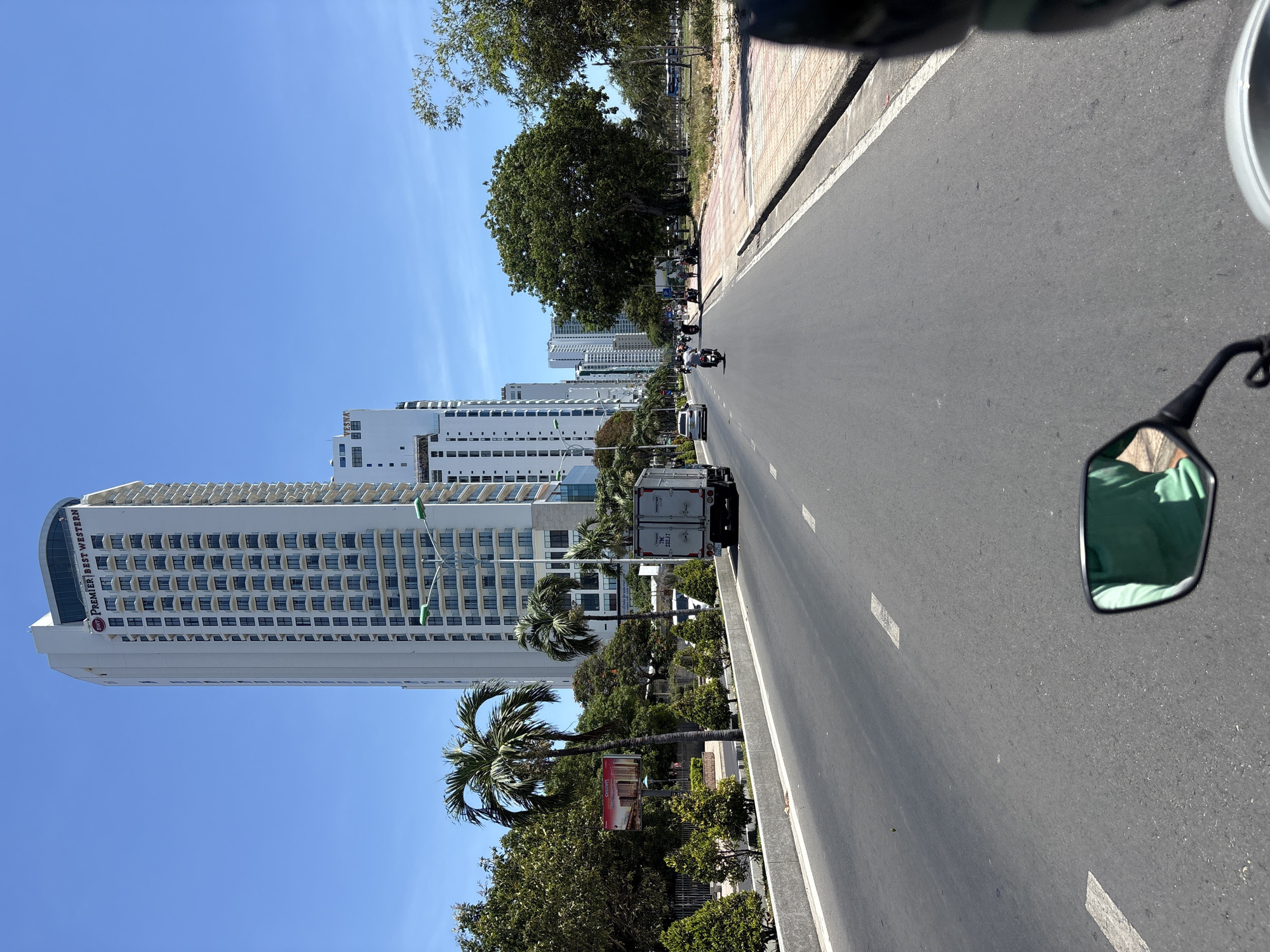 Captured from the perspective of a person riding a motorcycle on a road, the image portrays a bright day in Nha Trang Ward, Vietnam. A tall, white high-rise hotel building stands prominently on the left side of the road. Along the roadside, palm trees are interspersed with lush greenery.
Vehicles, including a truck and scooters, are visible on the road ahead. The sky is a clear, vibrant blue. The motorcycle's side mirror is visible on the right side of the frame, reflecting the rider's green shirt. The road is marked with white lane indicators. A sidewalk borders the road on the right, with more trees lining the edge.
