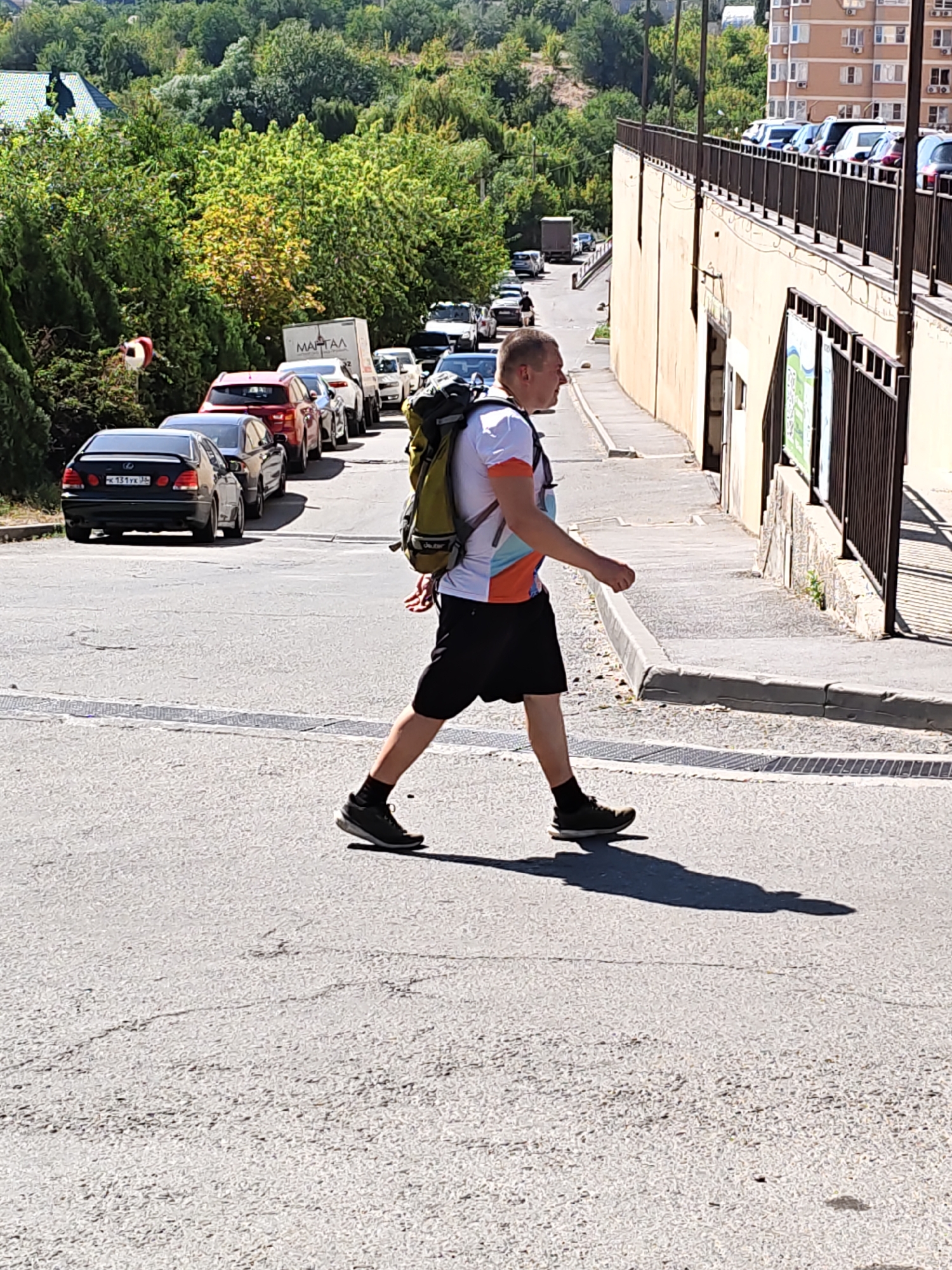 A man with short hair, wearing a white t-shirt with an orange and blue design on the sleeves, black shorts, black socks, and black athletic shoes, is walking across a paved road. He is carrying a large olive-green backpack. The man is walking from left to right in the frame, and his shadow is cast on the road behind him. In the background, a line of cars is parked on the side of the road. Further in the background, there are trees and buildings. The scene appears to be on a sunny day.
The location is on a street in Rostov-on-Don, Russia, specifically on the road leading up to the Rostov-on-Don embankment, in the vicinity of Gagarin Street. The architecture visible, including the multi-story apartment building on the right and the style of fencing, are consistent with this area. The presence of the cars and the pedestrian crossing further indicate an urban environment.
