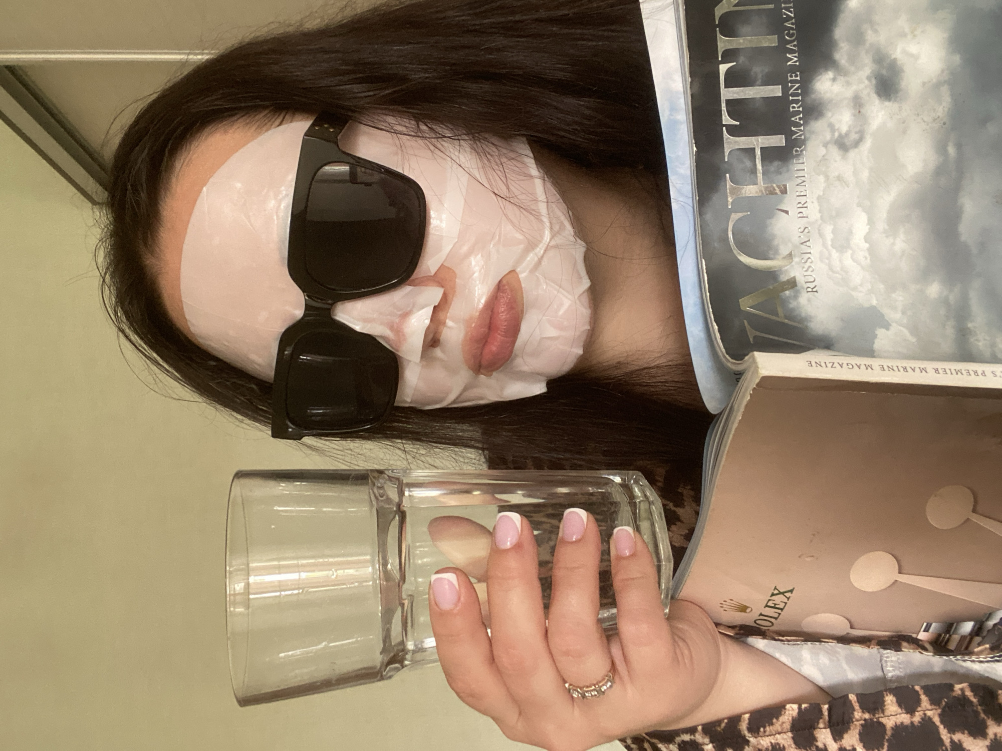 A woman is lying down wearing a white sheet mask and sunglasses. She is holding a glass of water with a French manicure on her nails. She is surrounded by magazines, one of which is titled "YACHTING: Russia's Premier Marine Magazine." The other magazine has "ROLEX" printed on its cover. Given the name of the magazine and its tagline, it can be inferred the picture was taken in Sochi, Russia. The woman is wearing a leopard-print shirt.
