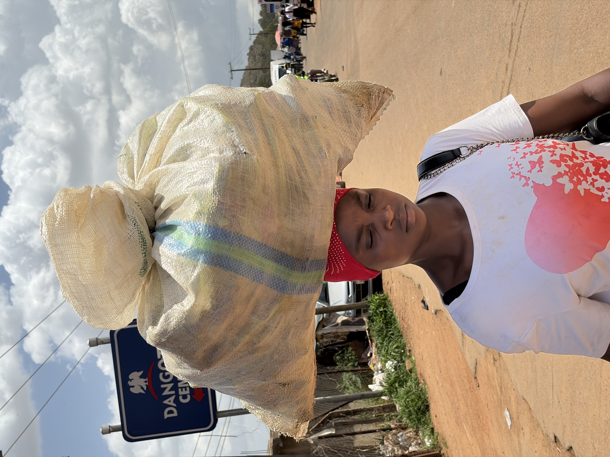 A close-up, rotated image captures a young Black woman skillfully balancing a large, heavy sack on her head while walking outdoors. The woman is positioned towards the right side of the frame. She wears a white t-shirt adorned with a red design featuring butterflies and a large red abstract shape on the lower front. A red headscarf or headband is visible around her forehead, and a black bag with a silver chain strap is slung across her body. Her eyes are closed or downcast, conveying focus or the effort of carrying her burden.
The voluminous sack, made of light-colored, coarsely woven material (possibly polypropylene or jute), is tied securely at the top and features blue and green stripes near its base. It dominates the left side of the frame, towering over the woman.
The setting appears to be a bright, daytime scene with a sky filled with large, fluffy cumulus clouds visible in the upper left. Thin power lines crisscross the cloudy sky. A reddish-brown dirt or unpaved road stretches into the background. On the left, partially obscured by the sack, is a blue rectangular sign displaying a white eagle emblem and the prominent text "DANGOTE CEMENT" with a downward-pointing arrow. In the distance, on the right side of the road, several indistinct figures of people and a white truck suggest a busy thoroughfare or market area. Sparse green vegetation lines the roadside. The scene captures a moment of daily life in Gura Top, Nigeria, highlighting a common method of transporting goods.