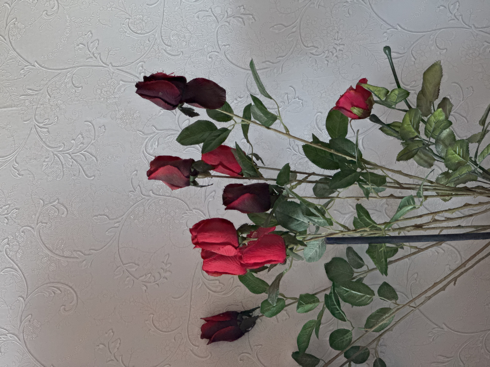 The image shows a bouquet of red roses lying on a surface with white textured wallpaper. The roses appear to be artificial, with varying shades of red and green leaves. The stems are visible, some with thorns. There is a dark, thin, horizontal object partially obscuring the stems. There are no people present. The location is Chaikovsky, Russia.