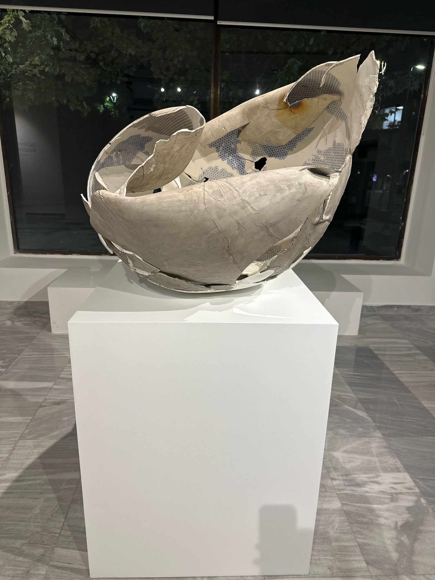 A large, abstract, ceramic sculpture, appearing to be broken or shattered into irregular pieces, is displayed on a white rectangular pedestal in what appears to be an art gallery. The sculpture has a rough, textured exterior in shades of gray and white, with hints of a blue and white patterned interior visible through the cracks and openings. The sculpture is lit from above, casting a shadow on the floor.
Behind the sculpture, a large window reveals an outdoor scene at dusk or night. Trees with visible branches and leaves are illuminated by ambient light. In the distance, streetlights and building lights are visible, suggesting an urban environment. The floor of the gallery is tiled with large, gray, marble-like slabs arranged in a geometric pattern. The overall impression is one of a contemporary art exhibition, possibly in Belgrade, Serbia, given the context. No people are visible in the image.