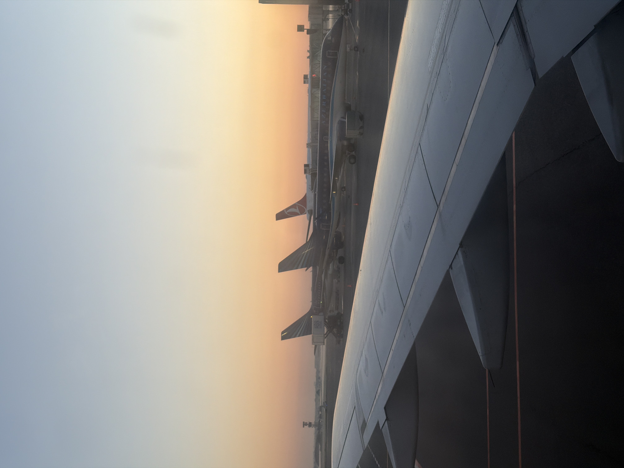 The image captures an airport tarmac at dawn or dusk, with the sky displaying a gradient of soft blues and oranges. In the foreground, a portion of an airplane's wing, viewed from inside the cabin, dominates the lower left and central frame. Beyond the wing, several passenger airplanes are parked at gates. Two of these aircraft, likely at a terminal, are closer and more detailed. One of them bears the livery and tail fin of Turkish Airlines, identifiable by its red and white logo. Another airplane, with "AZERBAIJAN" clearly visible on its fuselage, is also parked. In the background, various airport structures and equipment can be seen, including jet bridges connecting to the aircraft, and in the far distance, a control tower-like structure. The scene is likely at Heydar Aliyev International Airport (GYD), the main airport serving Baku, the capital of Azerbaijan, which is located in the Khazar Raion district of the city. There are no people visible in the image.