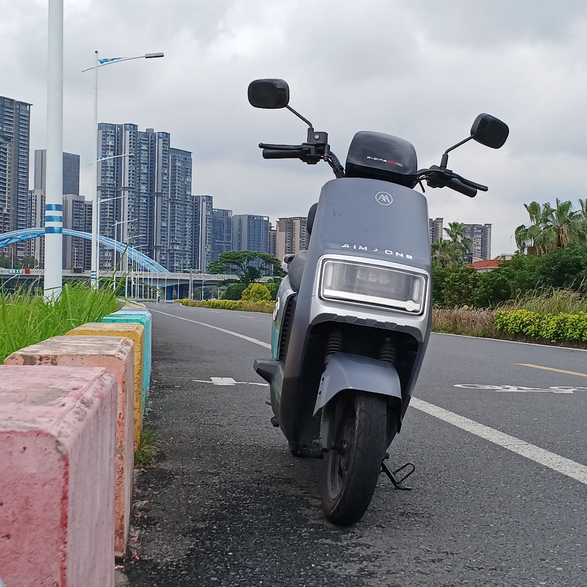 A gray AIM-ONE electric scooter stands parked on an asphalt road with a white bicycle lane marking in the Shunde District, China. To the left of the scooter, a low concrete barrier is painted with alternating sections of pink, orange, yellow, and blue. Tall grass grows between the barrier and the road. In the background, high-rise buildings dominate the skyline under a cloudy sky. A white lamppost with blue stripes and an arched blue bridge are visible among the buildings. Palm trees and other greenery are to the right of the scooter and the buildings in the distance. The scooter is facing the camera, with its handlebars, mirrors, and front headlight clearly visible.