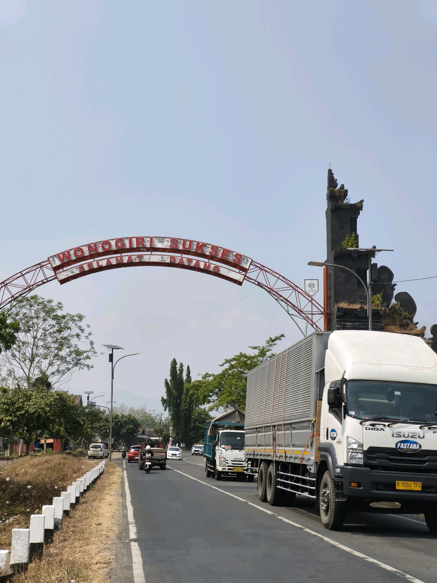 The image shows a street scene in Nambangan, Indonesia, under a clear blue sky. Dominating the background is a large red archway with white and red illuminated lettering that reads "WONOGIRI SUKSES SELAMAT DATANG." To the right of the archway is a tall, dark stone structure resembling a temple gate, adorned with greenery.
In the foreground, a white Isuzu truck is prominently featured, traveling on a gray asphalt road. The truck has the brand names "GIGA," "ISUZU," and "FASTANA LOGISTIC" visible. Its license plate is yellow with black text: "B 9204 TFZ." Another white truck is visible behind it.
The road is lined with white posts and dry grass on the left side. Other vehicles, including cars and motorcycles, are also present on the road, moving in the same direction as the trucks. Trees and some buildings can be seen on the left side of the road, and in the distance, mountains are faintly visible. The overall scene suggests a sunny day in a town or city, possibly a welcoming entrance.